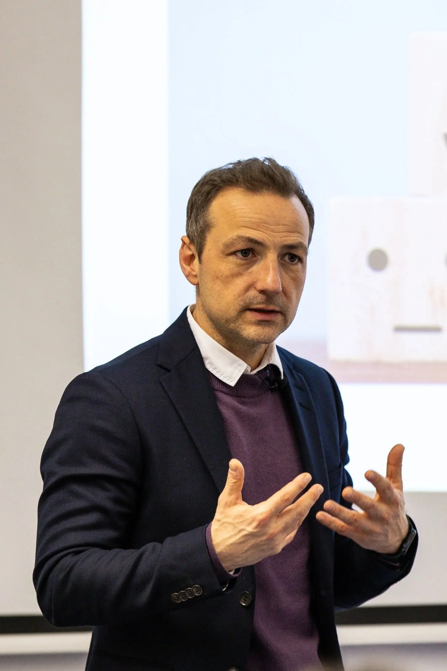 A man in a business suit gesturing with his hands during a presentation or discussion in a professional setting.