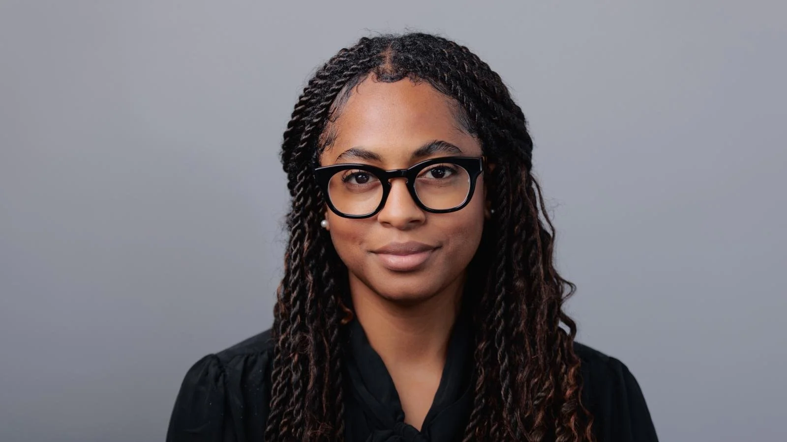 A woman with glasses and earrings, with long curly hair styled in twists, standing against a plain gray background.
