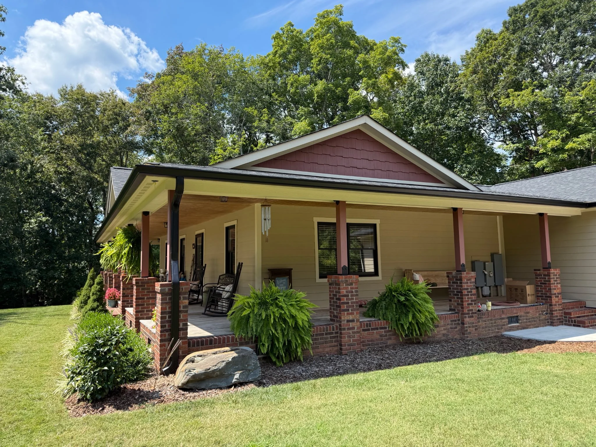 Back porch of a house with brick columns and lush green plants, overlooking a well-maintained yard with trees and a bright blue sky.
