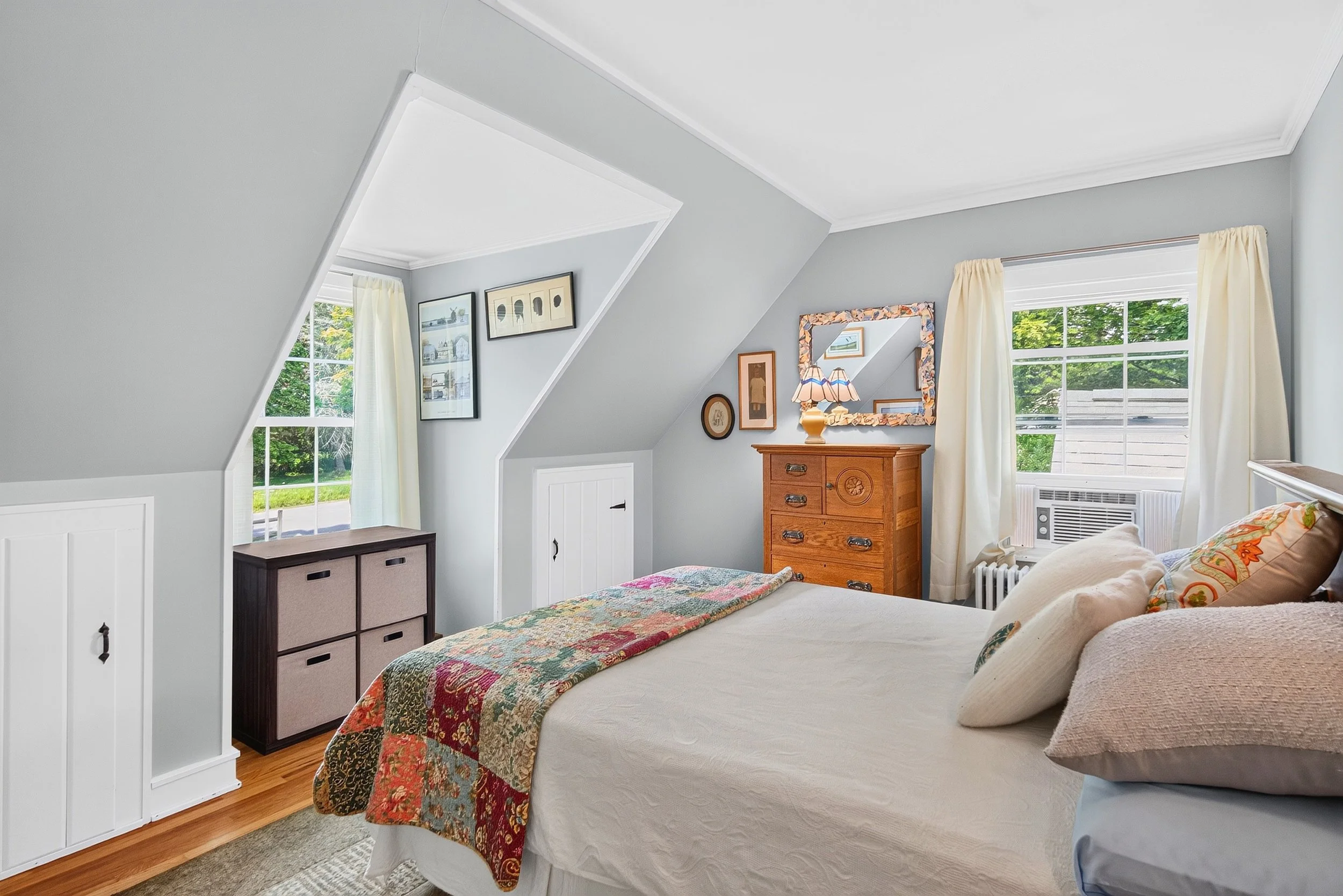 Interior real estate photography of a bright, cozy bedroom with sloped ceilings, large windows, and a neatly made bed