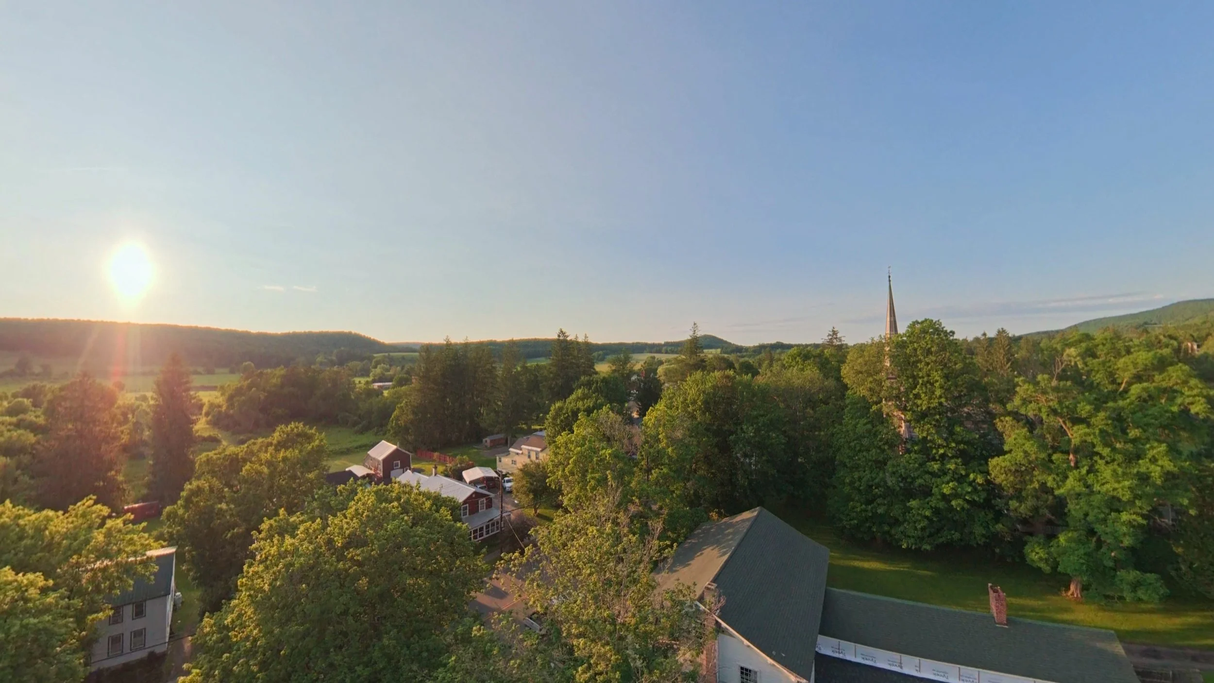 Professional aerial real estate photography showing a scenic neighborhood at sunset, with tree-lined properties, rooftops, and expansive countryside views captured by drone.