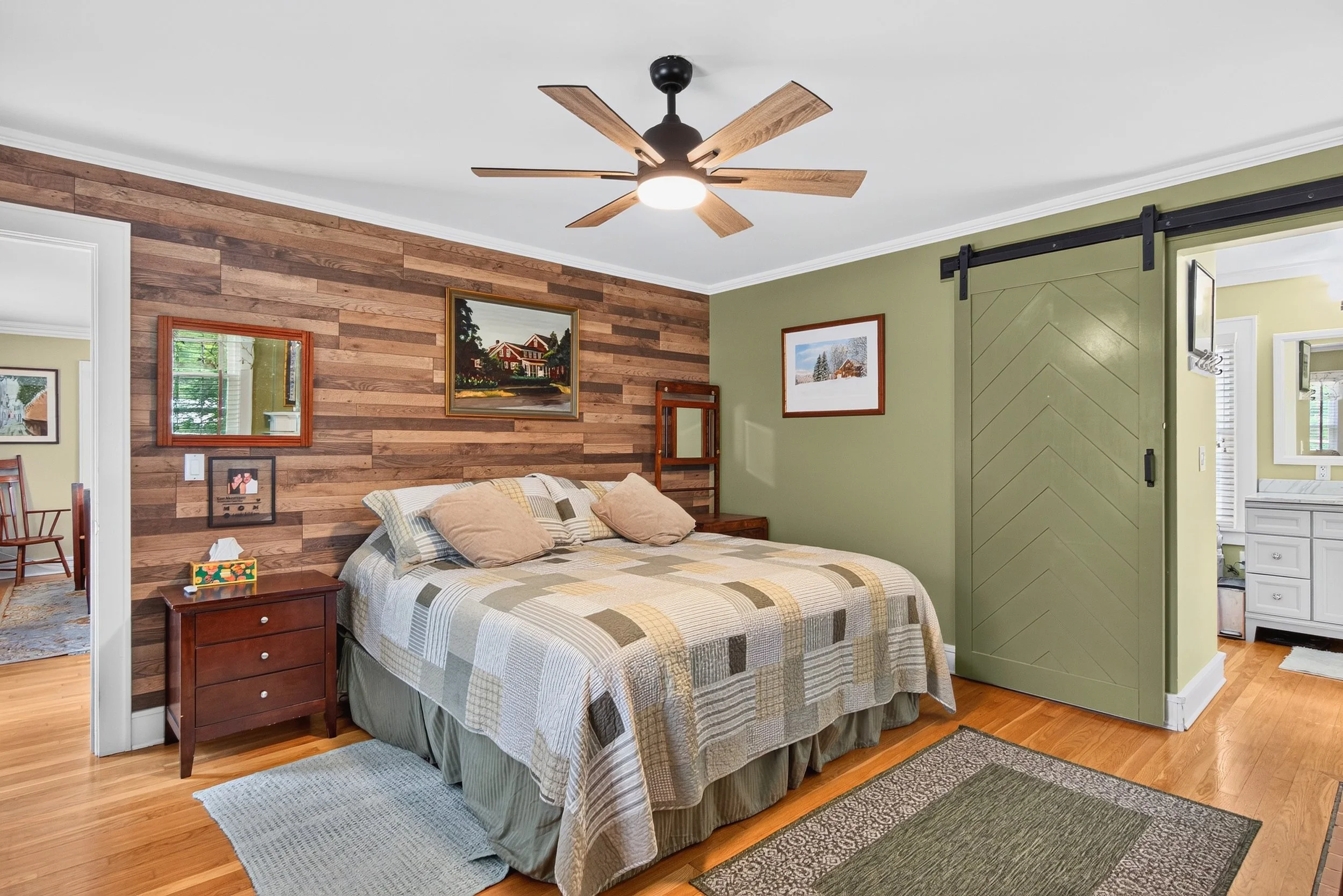 Interior real estate photography of a cozy bedroom featuring a rustic wood accent wall, ceiling fan, sliding barn door, and warm natural finishes.