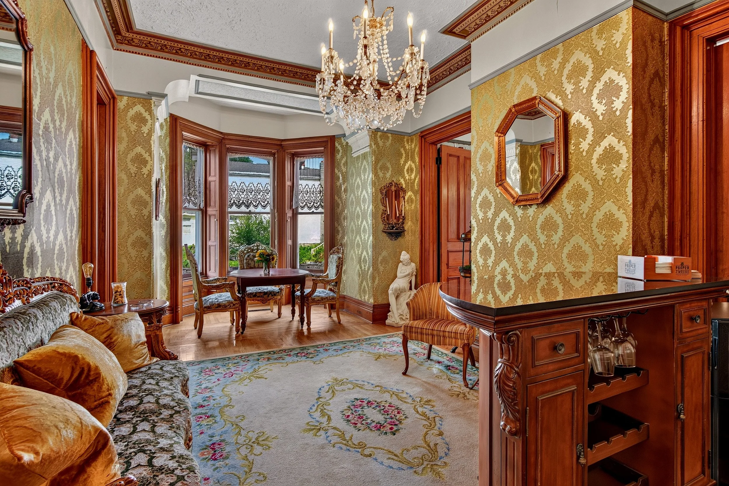 Interior real estate photography of an elegant historic home featuring a crystal chandelier, ornate wallpaper, decorative furnishings, and a sunlit bay window seating area.