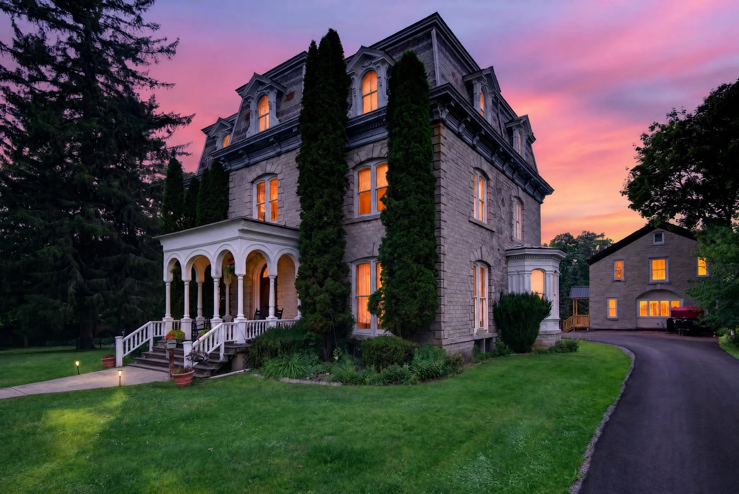 Twilight real estate photography of a historic stone mansion with illuminated windows, an elegant front porch, and a colorful sunset sky.
