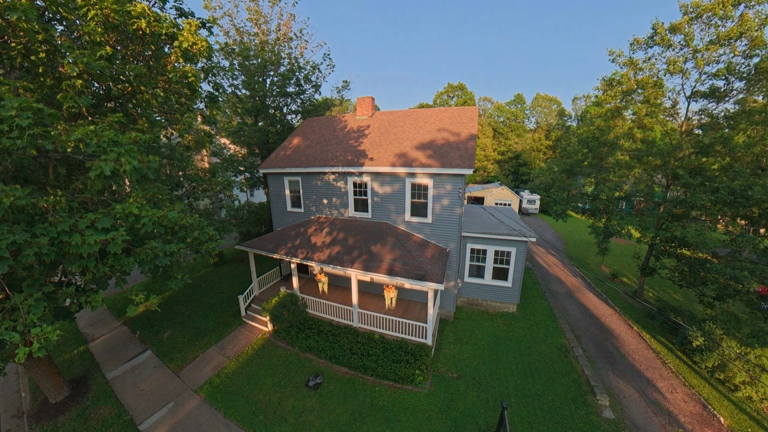 Professional aerial real estate photography of a two-story home with a red roof, covered front porch, green lawn, and mature trees, captured from an elevated drone perspective.