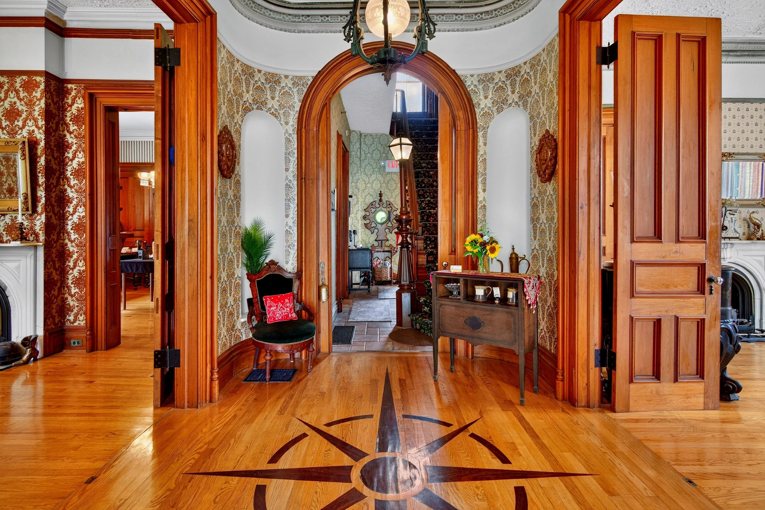 Interior real estate photography of an elegant historic home entryway featuring an arched wooden doorway, decorative hardwood floors, antique furnishings, and a chandelier.
