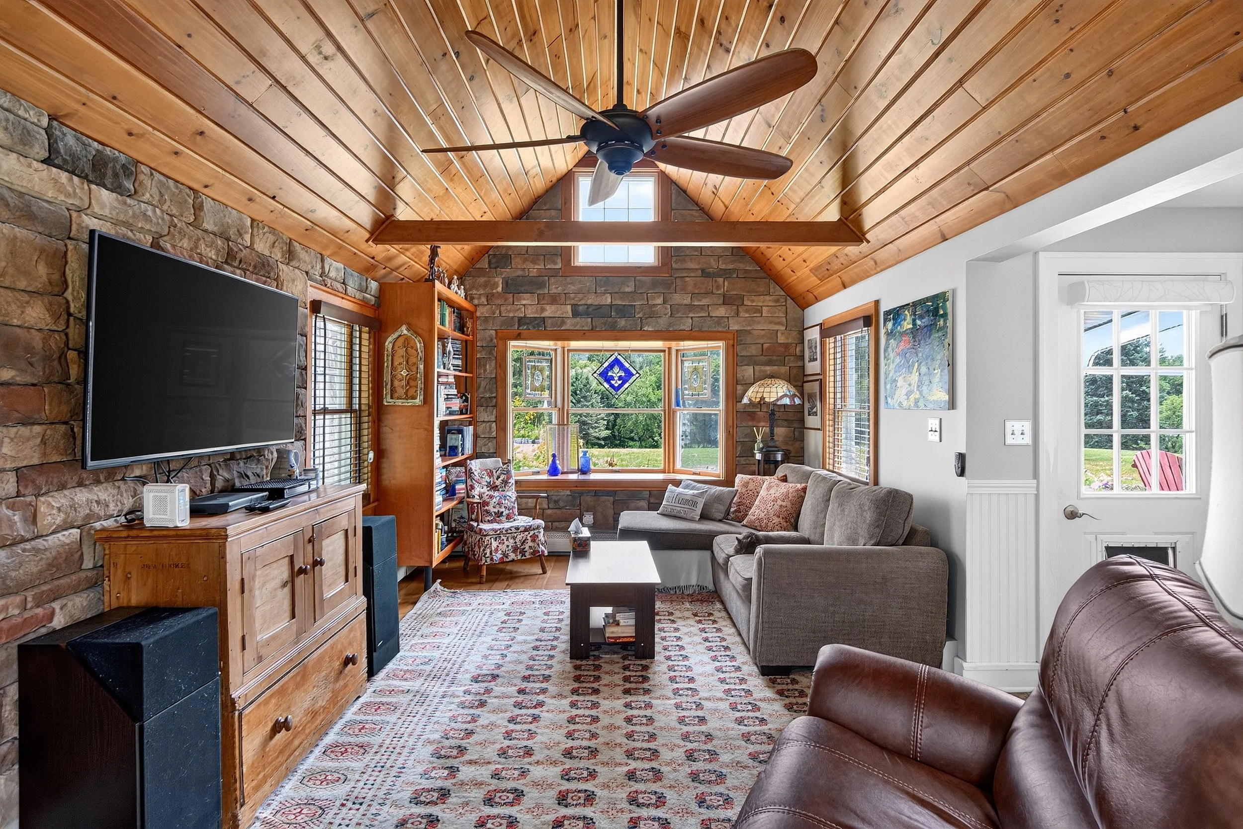 Professional real estate photography of a cozy living room with vaulted wooden ceilings, exposed stone walls and large windows.