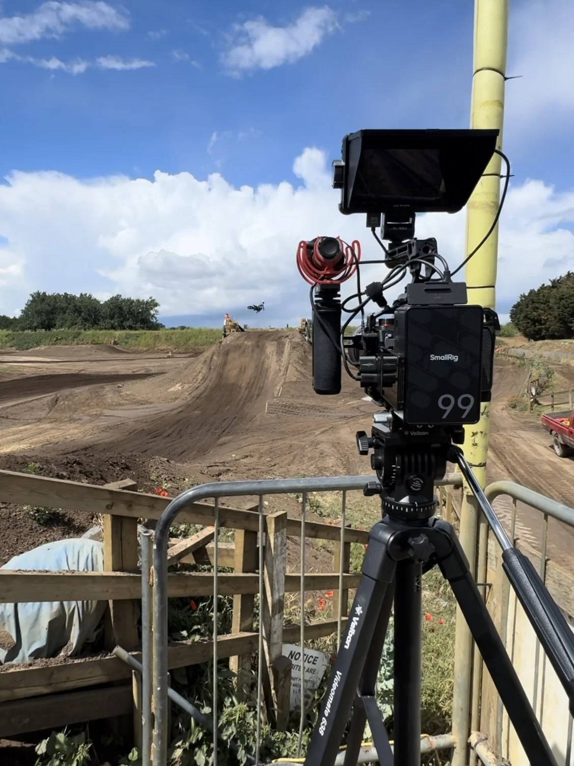 Camera setup filming a dirt bike jump at an outdoor motocross track on a sunny day with blue sky and clouds.