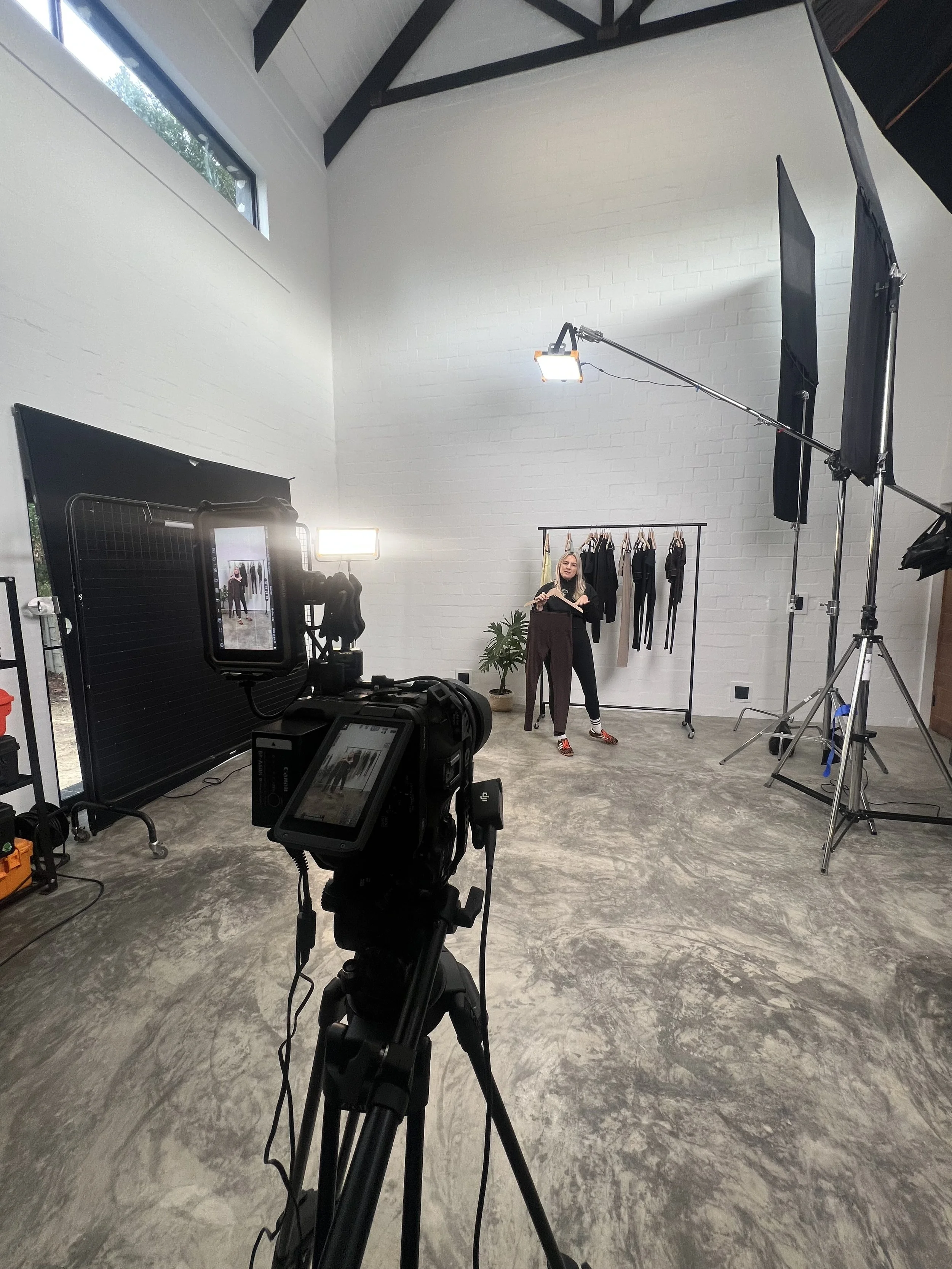 A behind-the-scenes view of a photography or filming studio with professional lighting equipment, camera, and a woman standing in front of a clothing rack with various garments. The studio has high white brick walls, a high ceiling with exposed beams, and a large window letting in natural light.