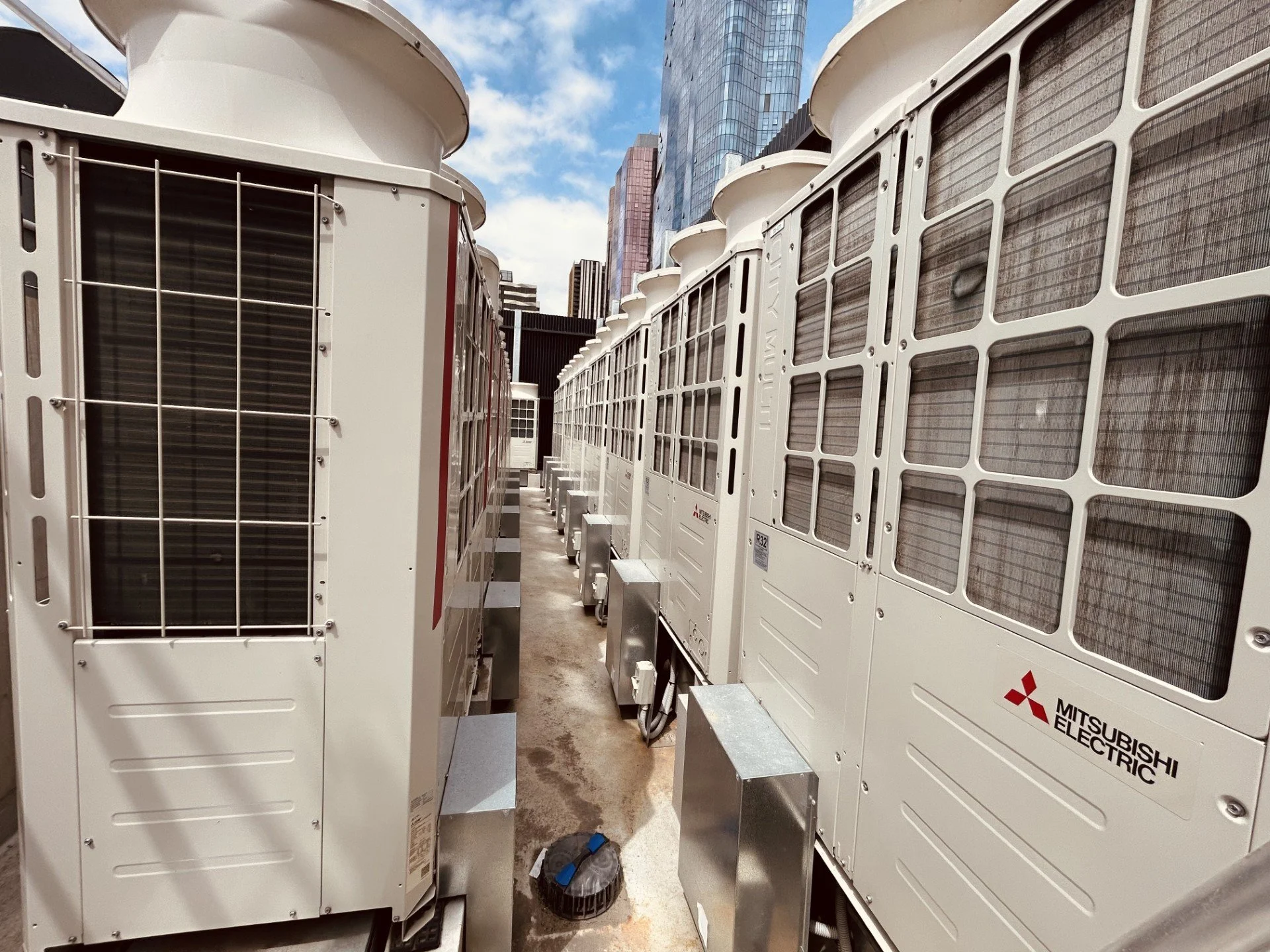 Multiple Mitsubishi Electric air conditioning units installed on a rooftop against a backdrop of tall modern skyscrapers and a partly cloudy sky.