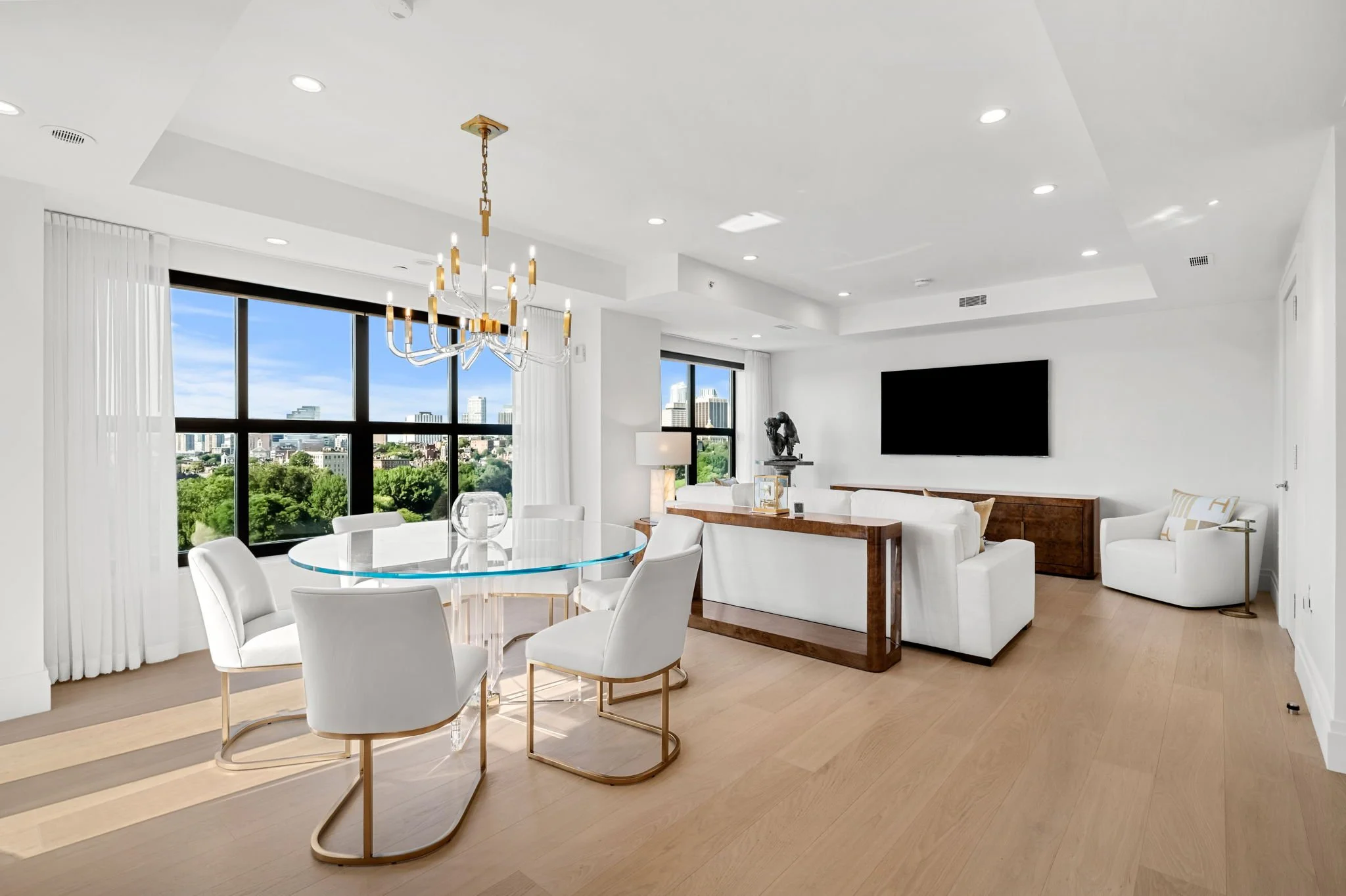 Modern apartment living room with large windows, white furniture, a glass dining table with white chairs, a chandelier, and cityscape view.
