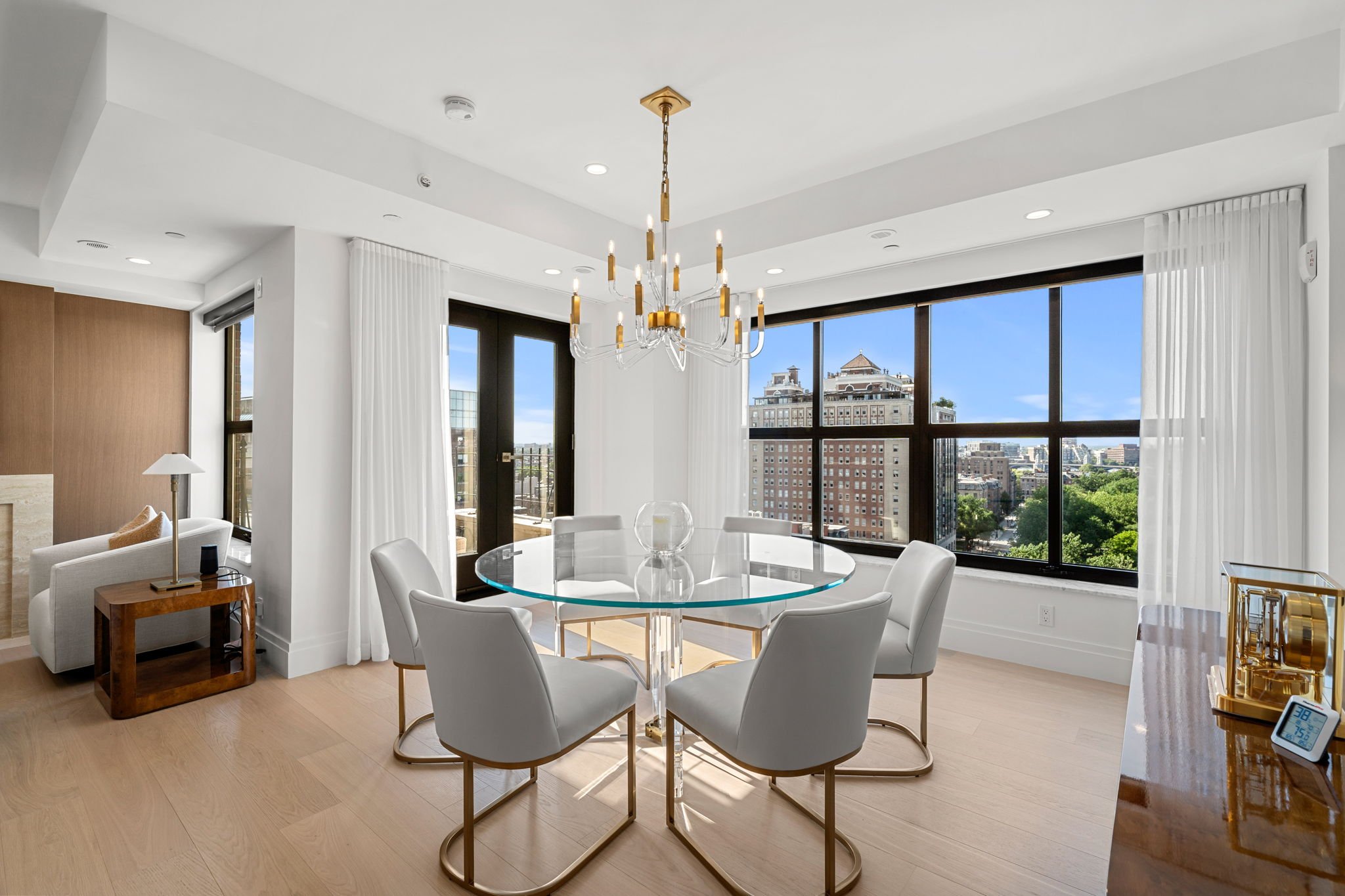 Modern dining room with a round glass table, white chairs, and large windows showing a cityscape and trees, with a brass chandelier hanging from the ceiling.