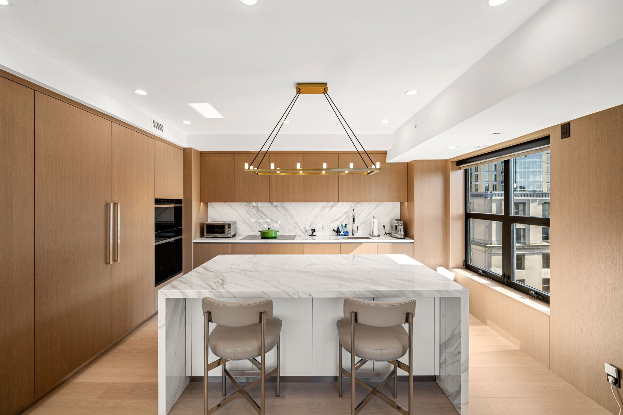 Modern kitchen with wooden cabinets, white marble island, and large window showing city buildings.