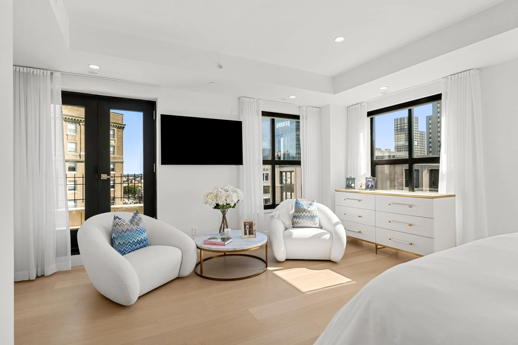 Modern living room with white walls, large windows, white furniture with colorful pillows, a TV on the wall, and city buildings visible outside.