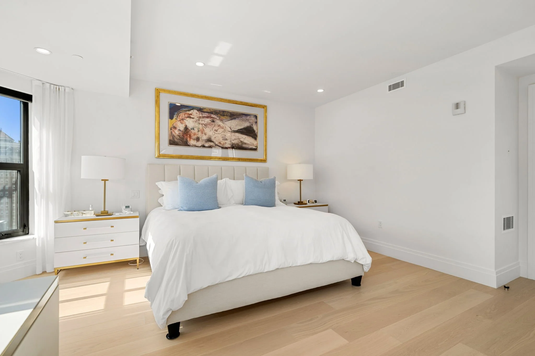 A bedroom with a neatly made bed with white bedding and two blue pillows. There are two white nightstands with gold accents, each topped with a white lamp. A large abstract painting with a gold frame hangs above the bed. A window with white curtains 