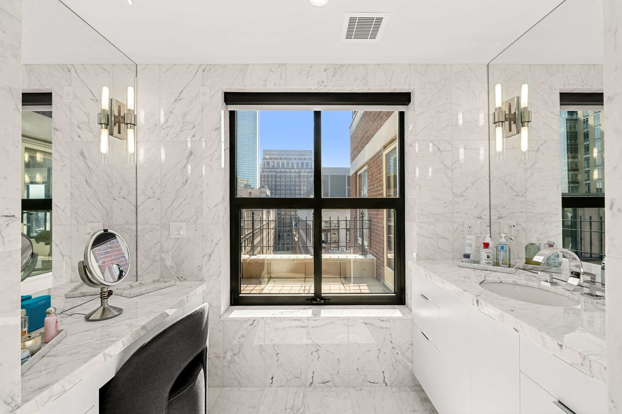 A bathroom with white marble walls and counters, a large window showing a cityscape with tall buildings, wall-mounted light fixtures, and various toiletries on the counter.