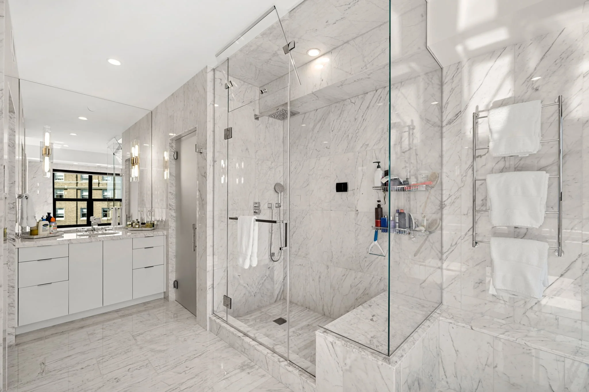 A modern bathroom with marble walls and floors, a glass shower enclosure, a white vanity with storage drawers, and a mirror with lighting fixtures.