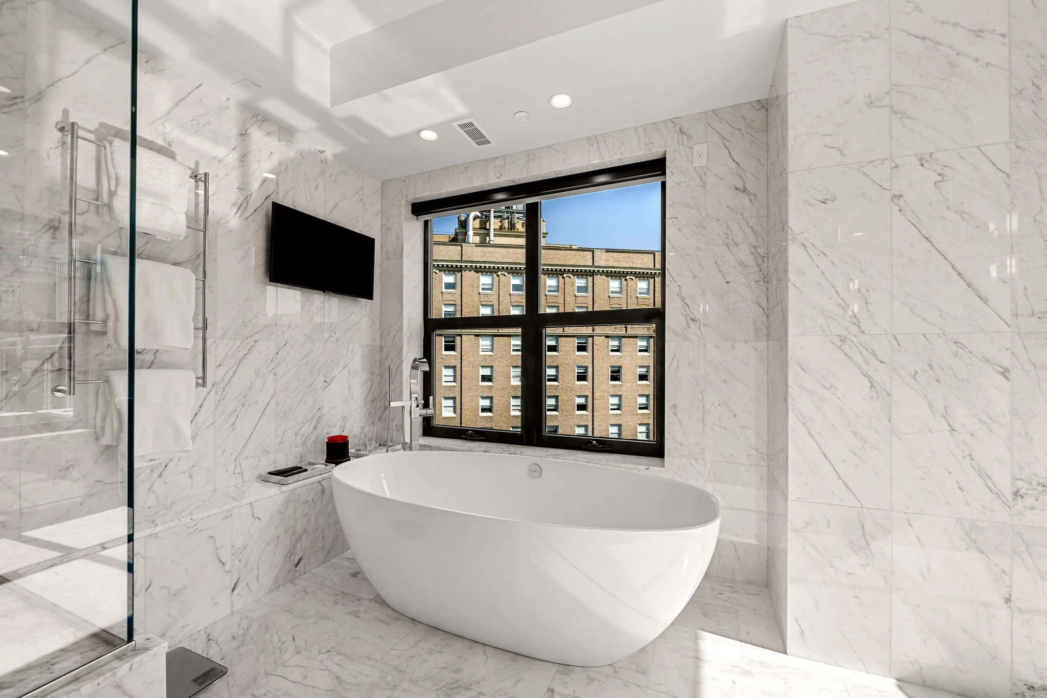 Modern bathroom with white marble walls and floor, a freestanding bathtub, a black window showing a cityscape, a wall-mounted TV, and towel storage.