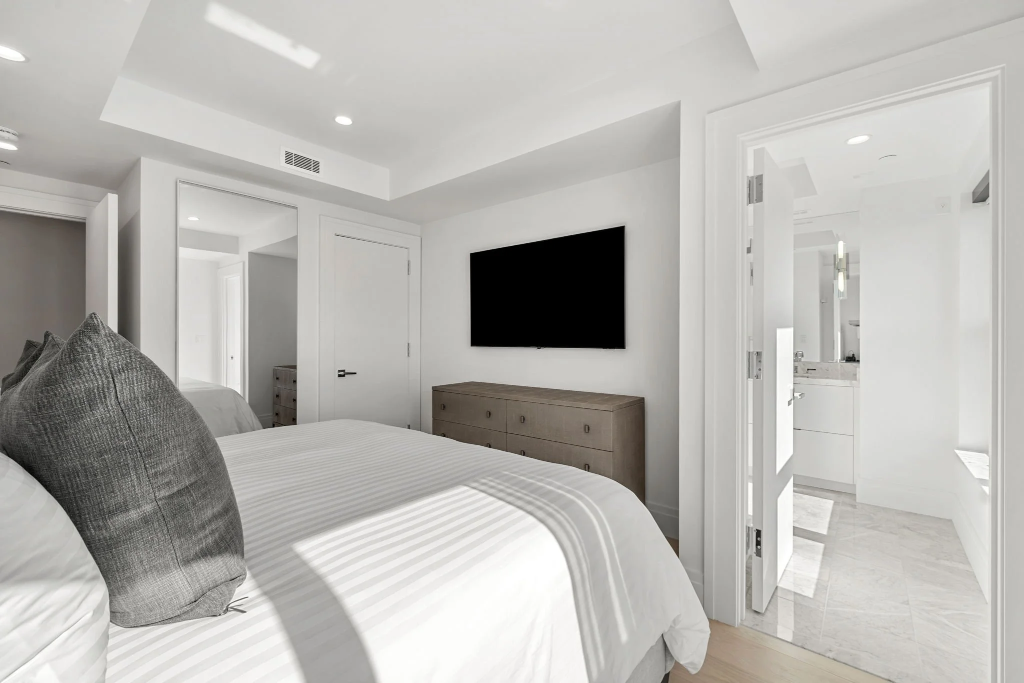 A modern, minimalist bedroom with a flat-screen TV mounted on the wall, a wooden dresser, a bed with gray pillows, and an open door leading to a bathroom with a marble floor and a vanity with two sinks.