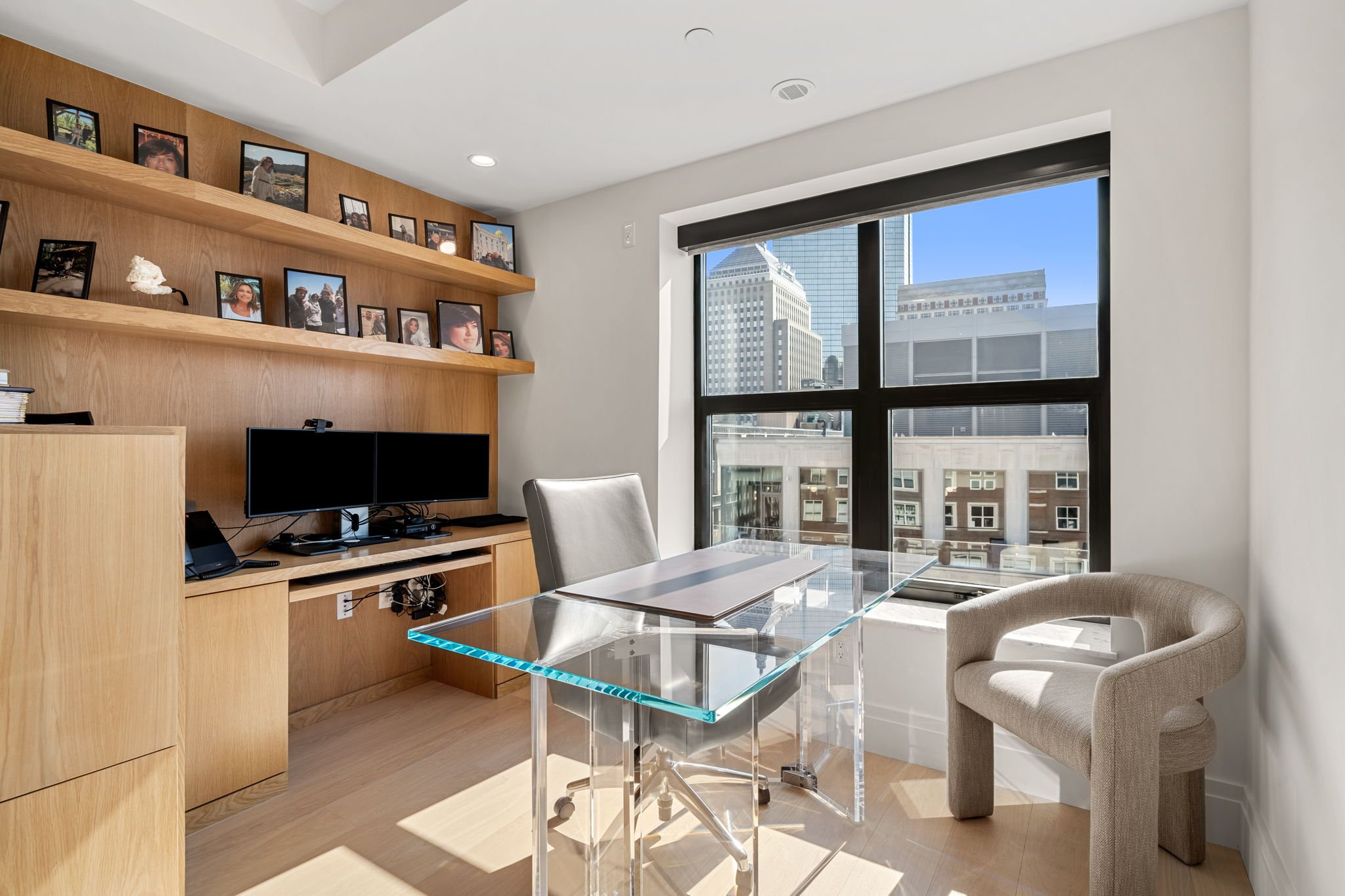 Modern office with large window showing city buildings, a glass desk with papers, a beige armchair, wooden shelves with framed photos, and electronic devices.