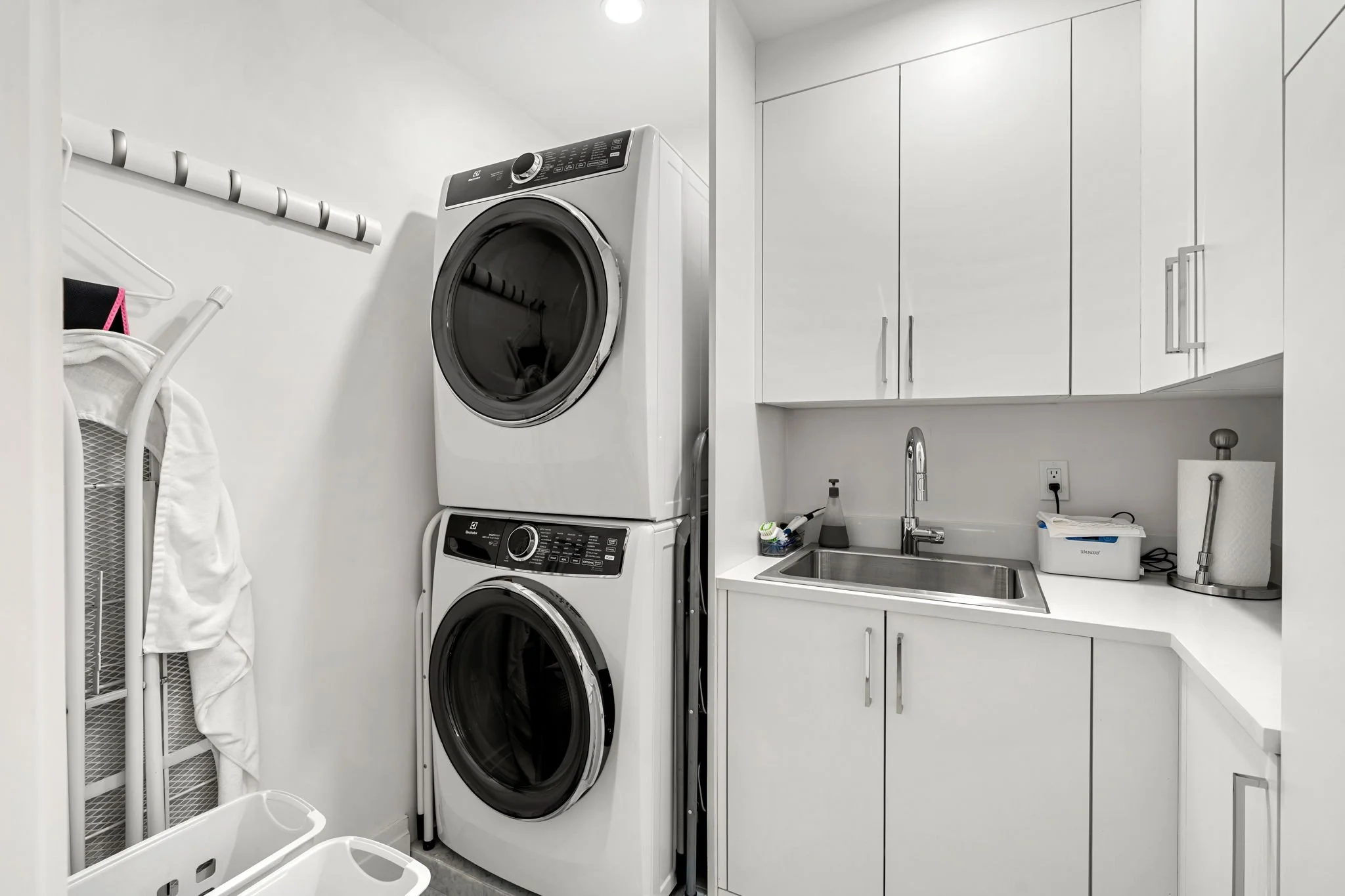 A modern laundry room with white cabinets, a stacked washer and dryer, a utility sink, and laundry supplies.