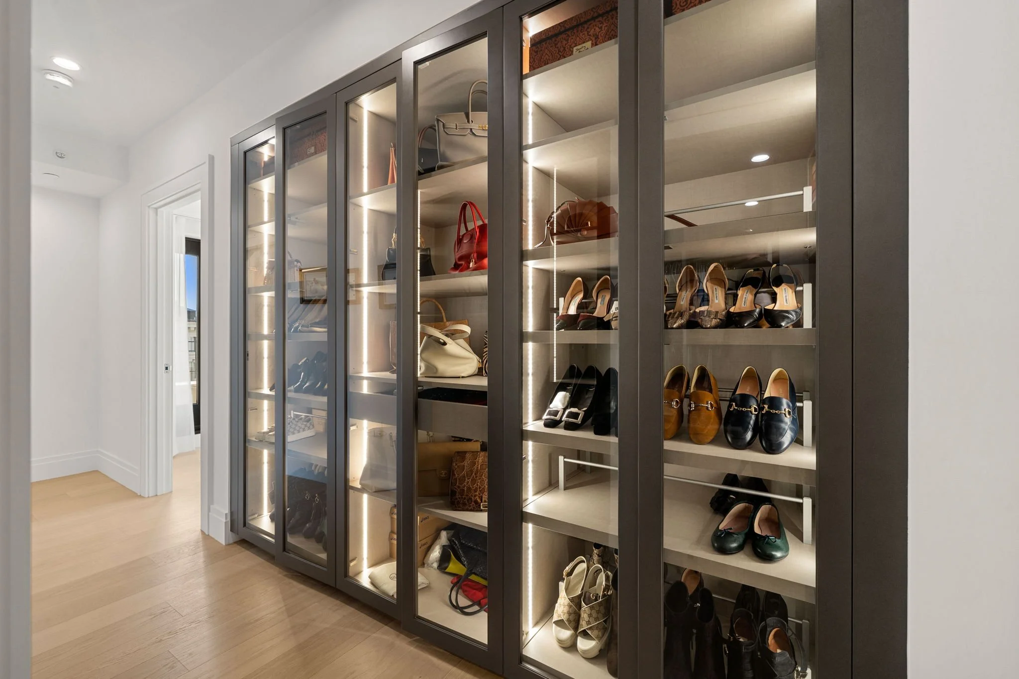 A glass-front shoe and handbag display cabinet with interior lighting, containing various shoes and handbags, situated in a modern room with white walls and wooden flooring.