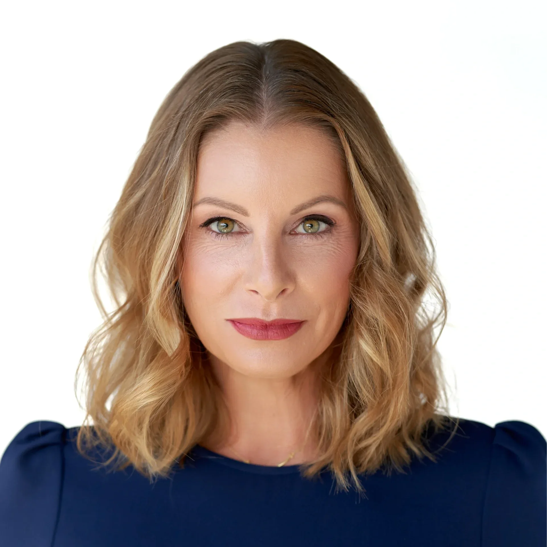 Close-up of a woman with wavy blonde hair, green eyes, and a friendly smile, wearing a dark blue top, against a plain white background.
