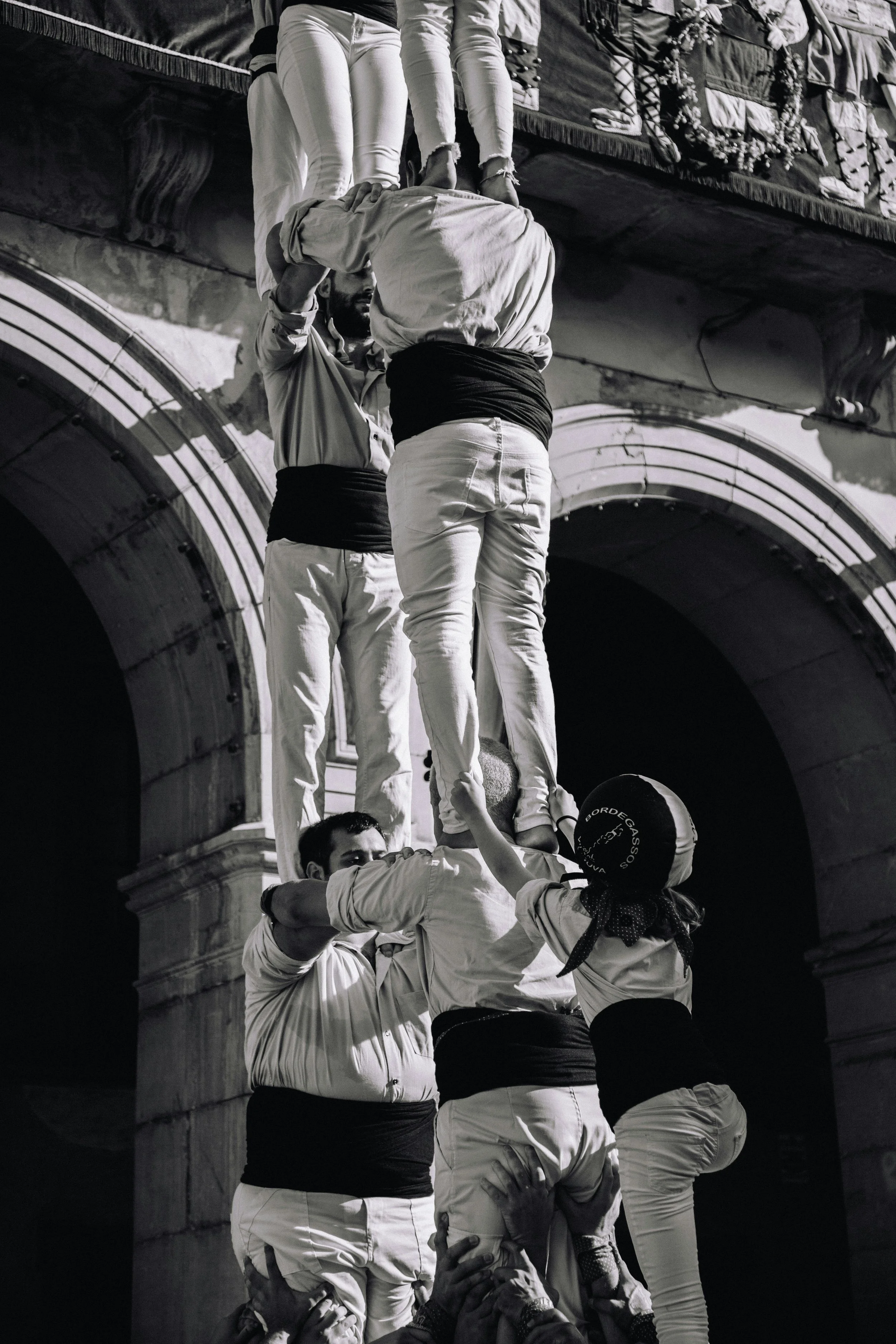 Black and white photo of people forming a human tower, with two people standing on the shoulders of others, holding each other's hands for balance, in front of historic architecture.