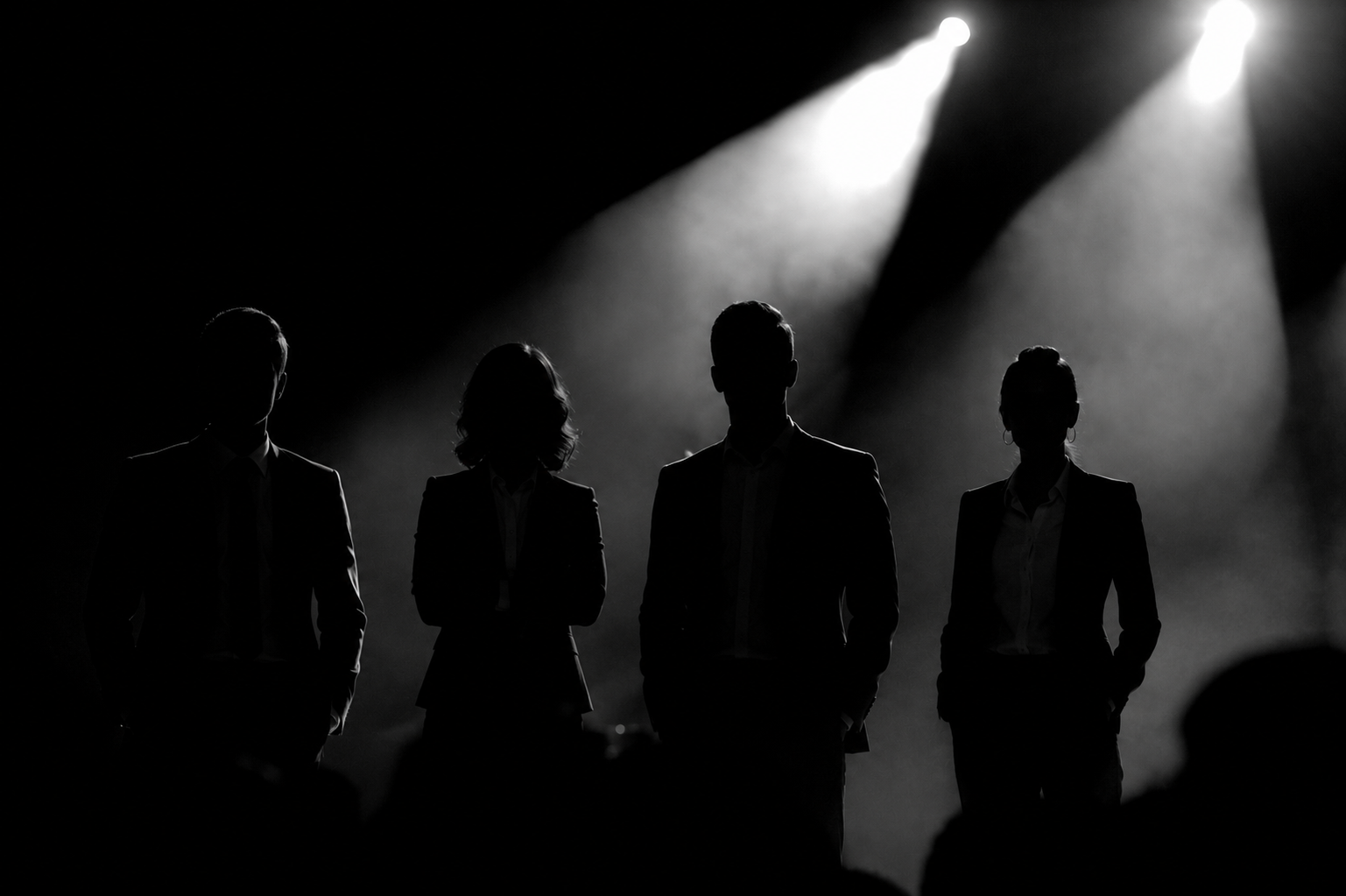 Silhouettes of four people standing on a stage with bright spotlights shining down from above, creating a dramatic, shadowed effect.