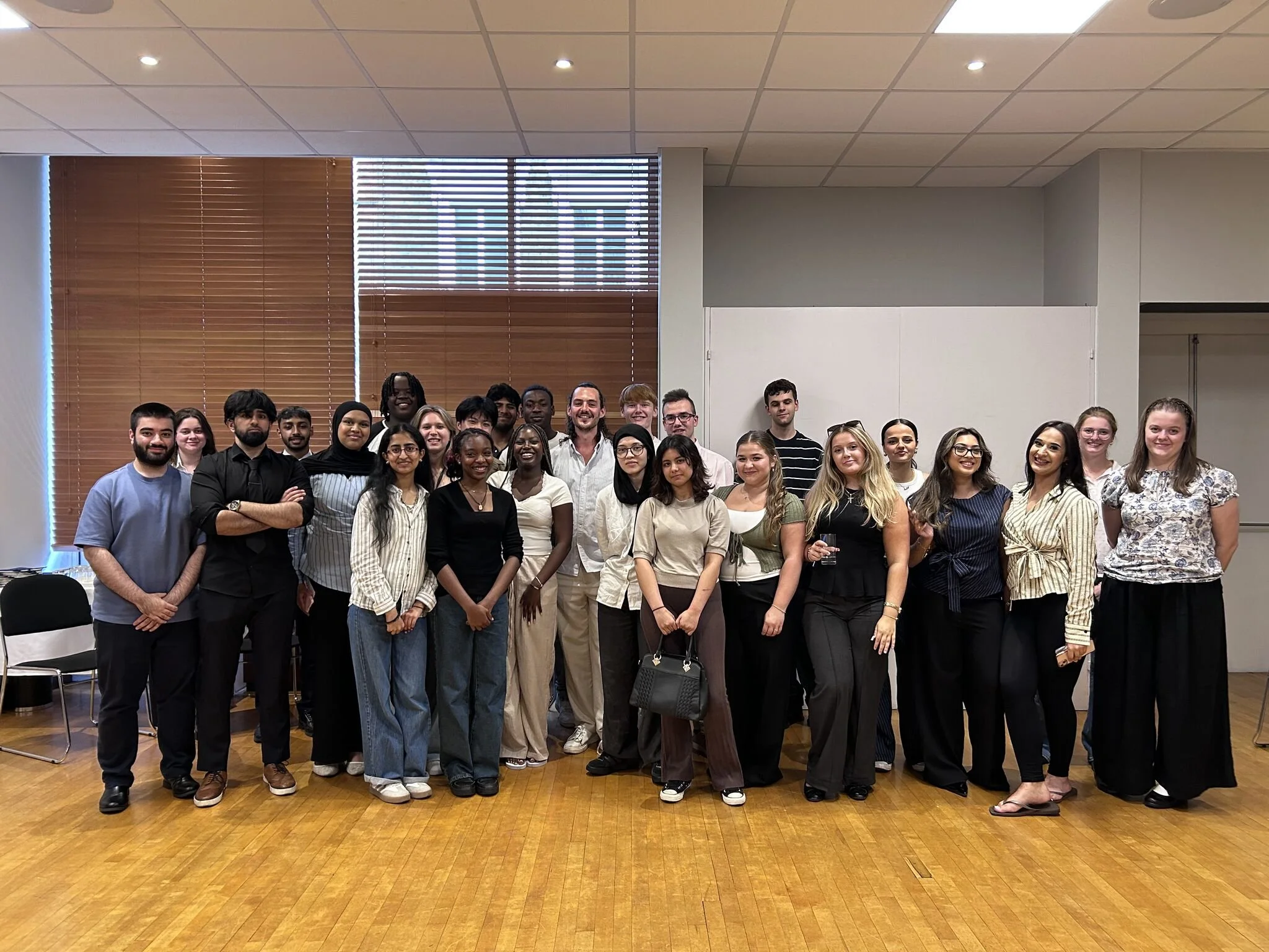 A diverse group of people standing together in a room with wooden floors, white walls, and horizontal wooden blinds on the windows, smiling at the camera.