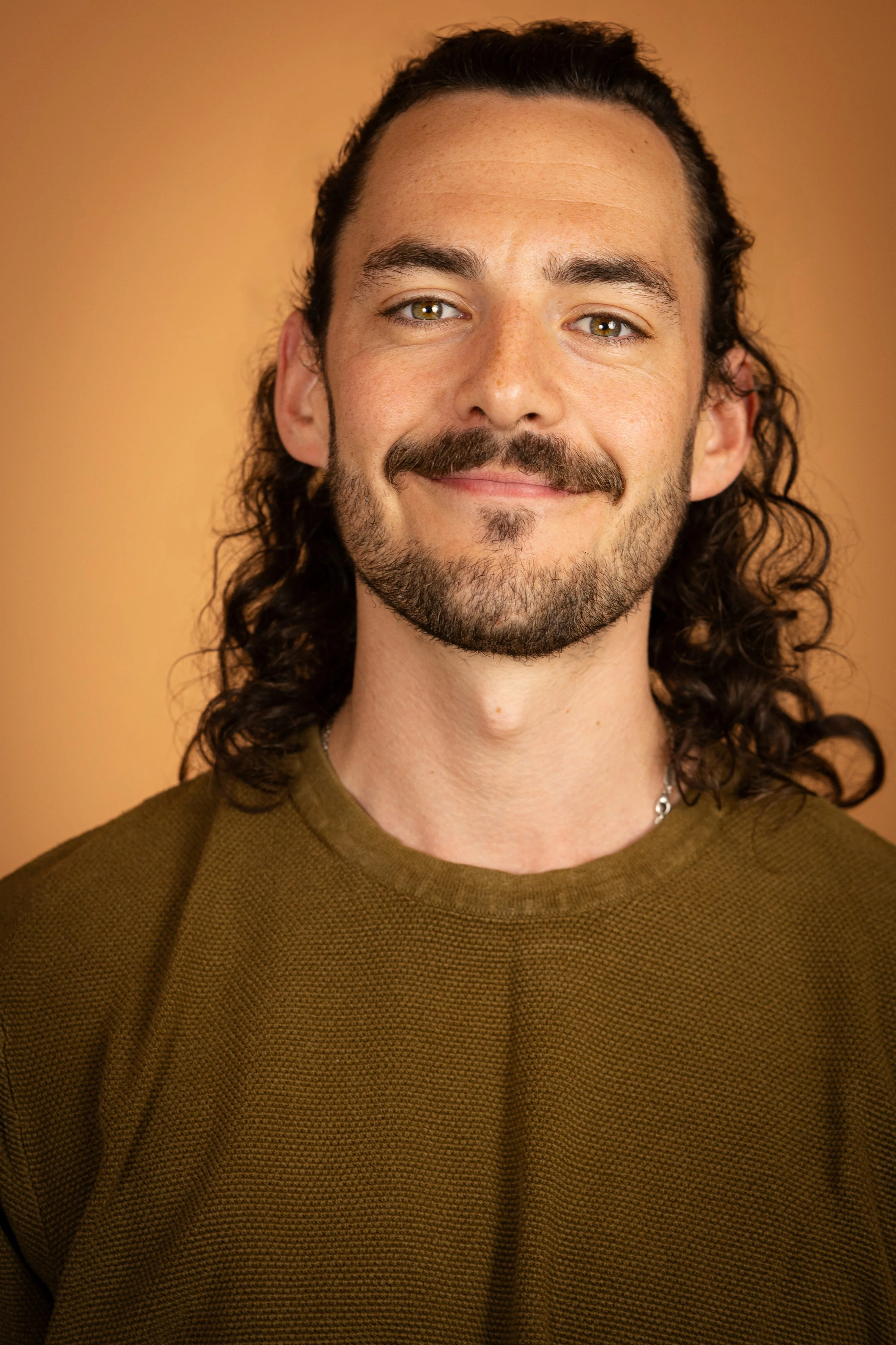Close-up of a man with long curly hair, a mustache, and a beard, smiling slightly, wearing a brown textured sweater, with a warm beige background.