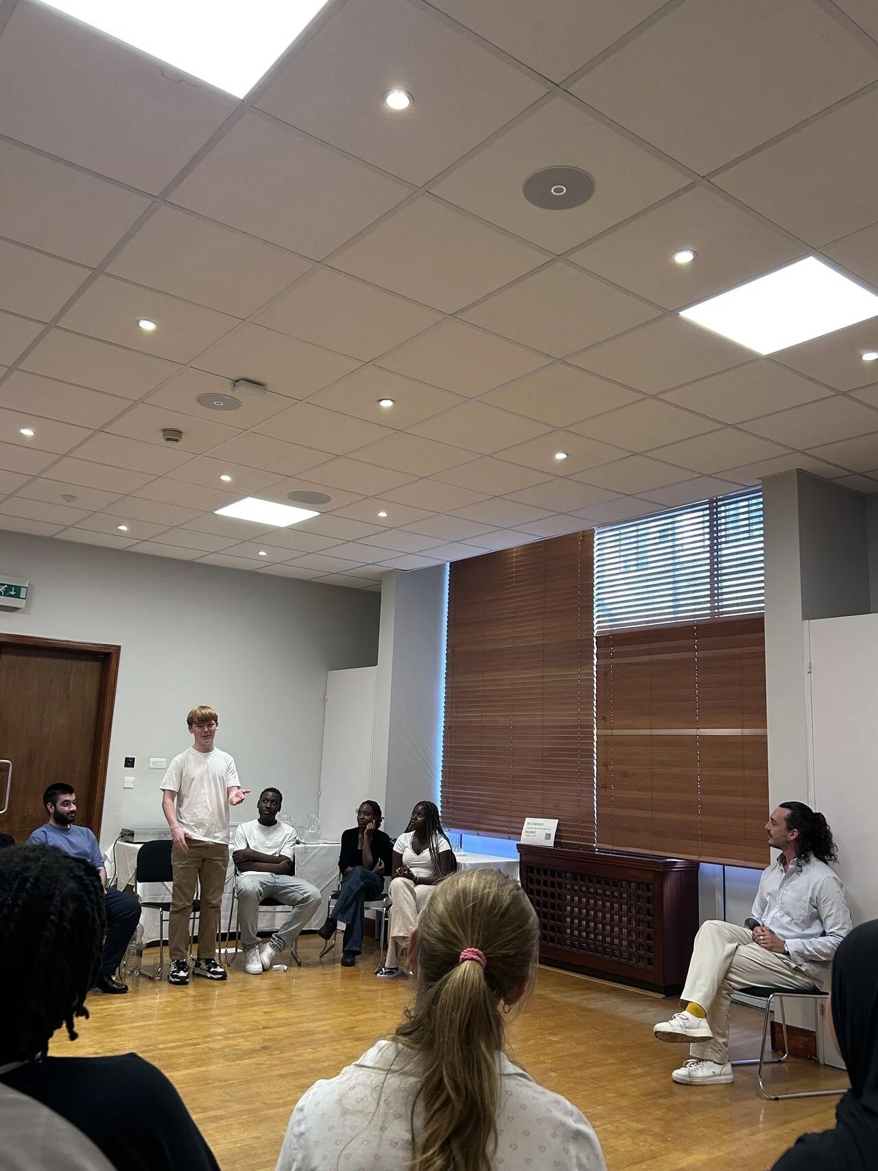 People participating in a discussion or presentation in a room with large windows and wooden blinds, some seated and one standing speaking.