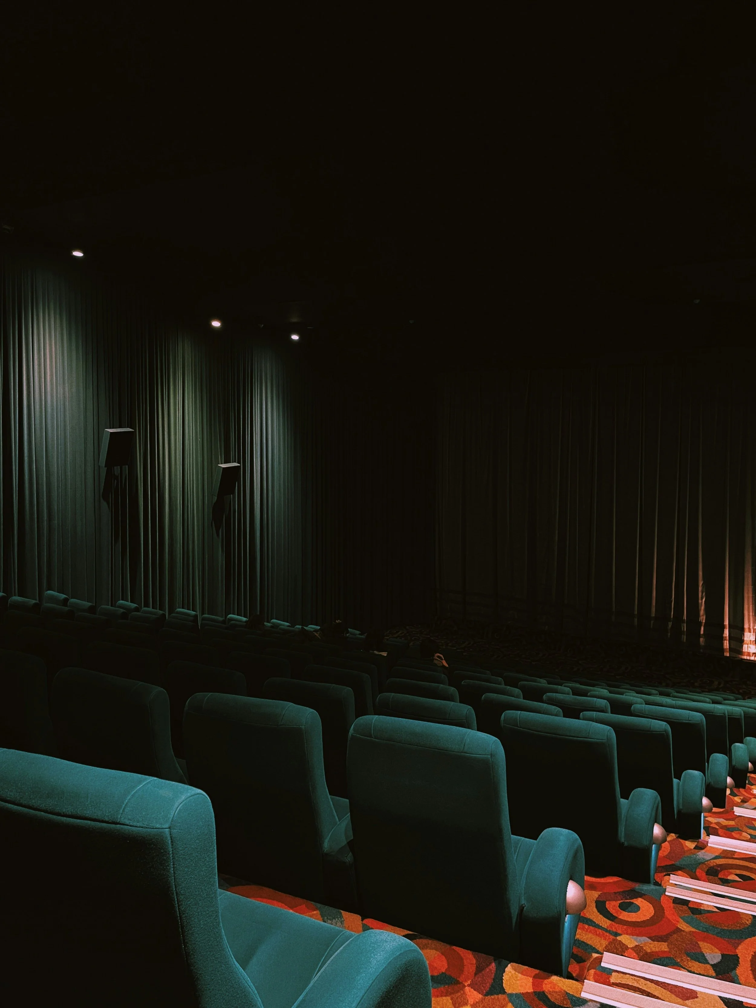 An empty movie theater with teal seats, a multicolored patterned carpet, closed dark curtains, and dim lighting.