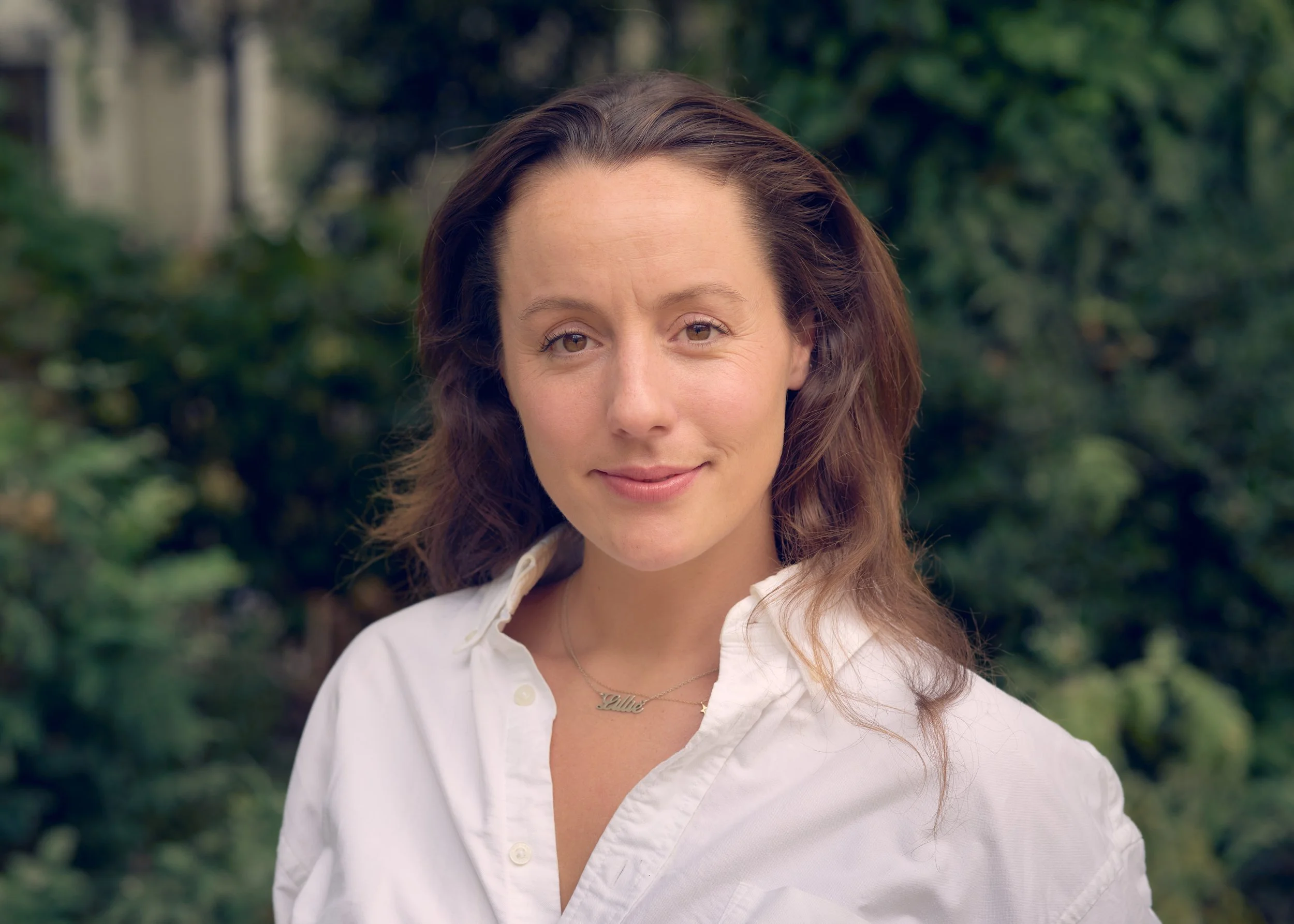 A woman with brown, wavy hair and light skin, wearing a white collared shirt and a name necklace that says "Lille," standing outdoors in front of blurred green foliage.