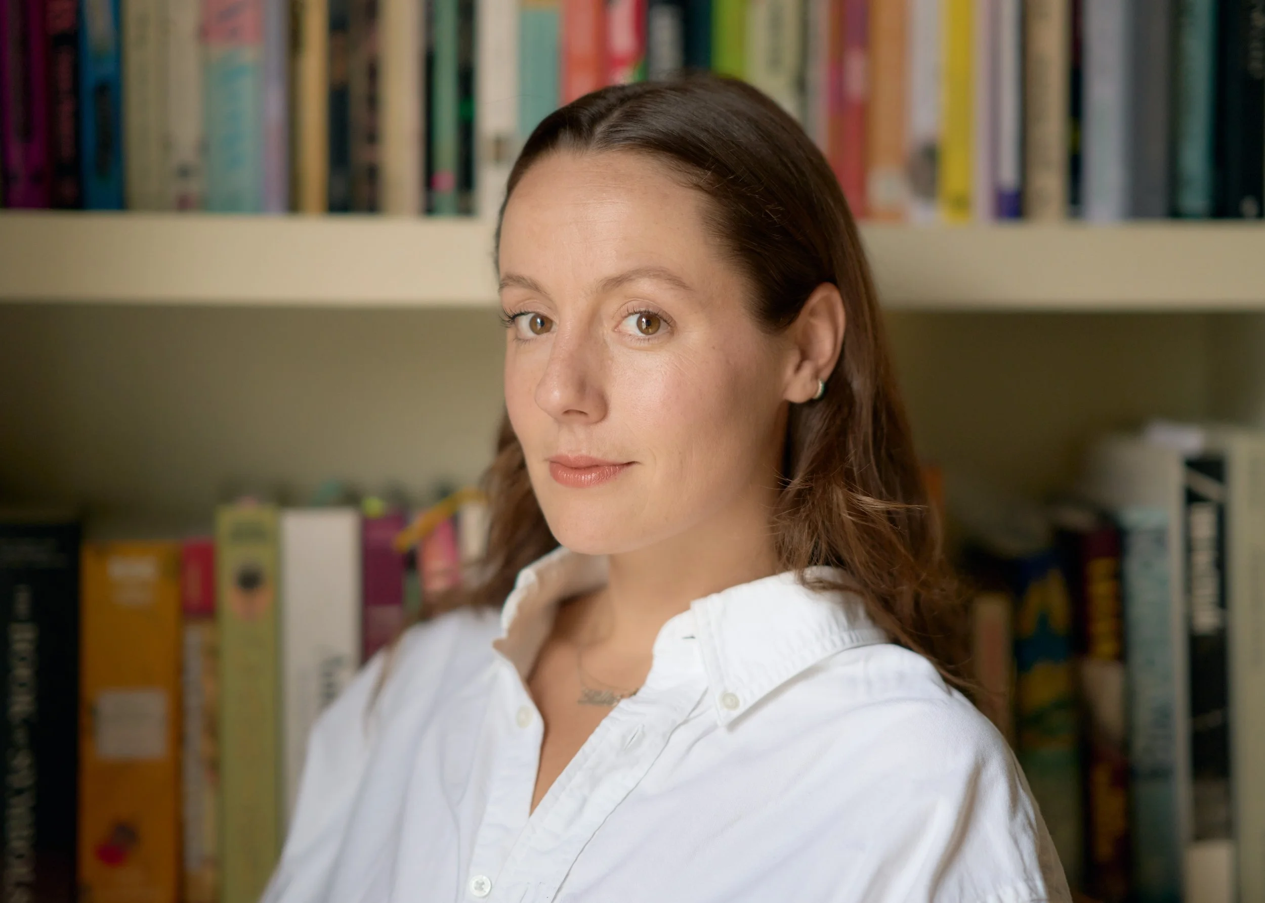 A woman with brown hair and hazel eyes wearing a white shirt in a room with a bookshelf filled with books.