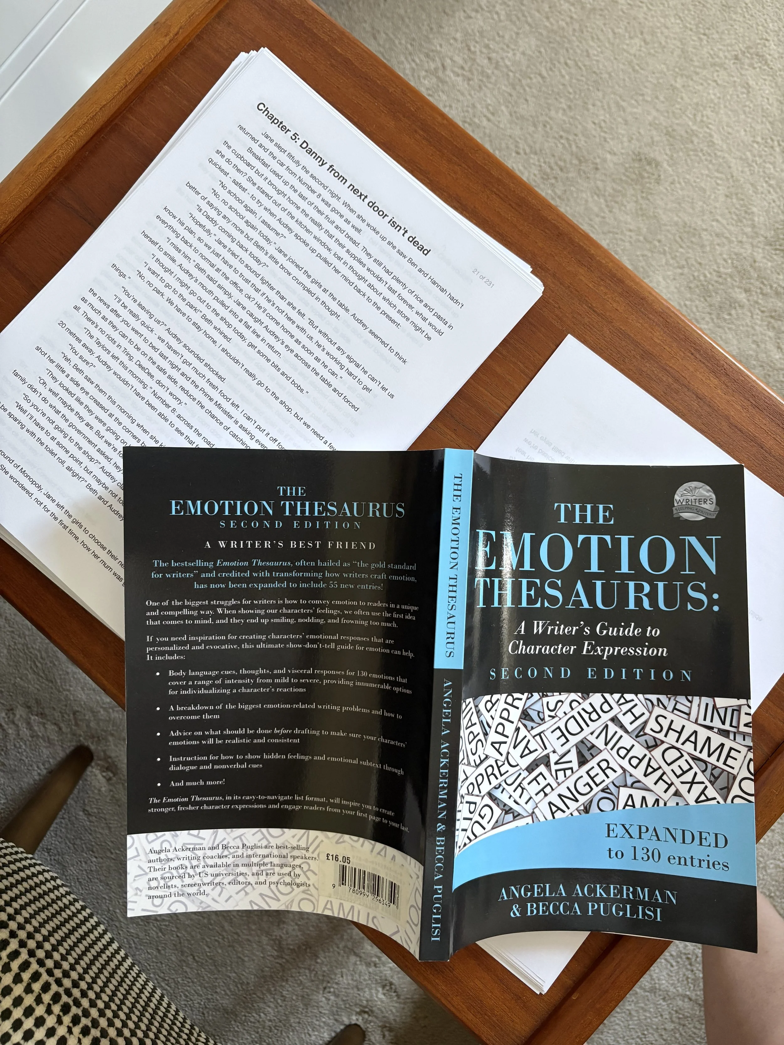 Photo of a manuscript on a table with the Emotion Thesaurus open on top of it, face down so you can see the front cover.