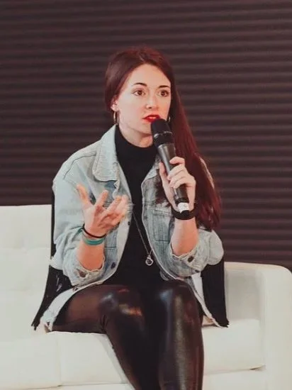 Amie wearing all black with a pale blue denim jacket speaking into a microphone on a panel at Blogosphere in 2019.