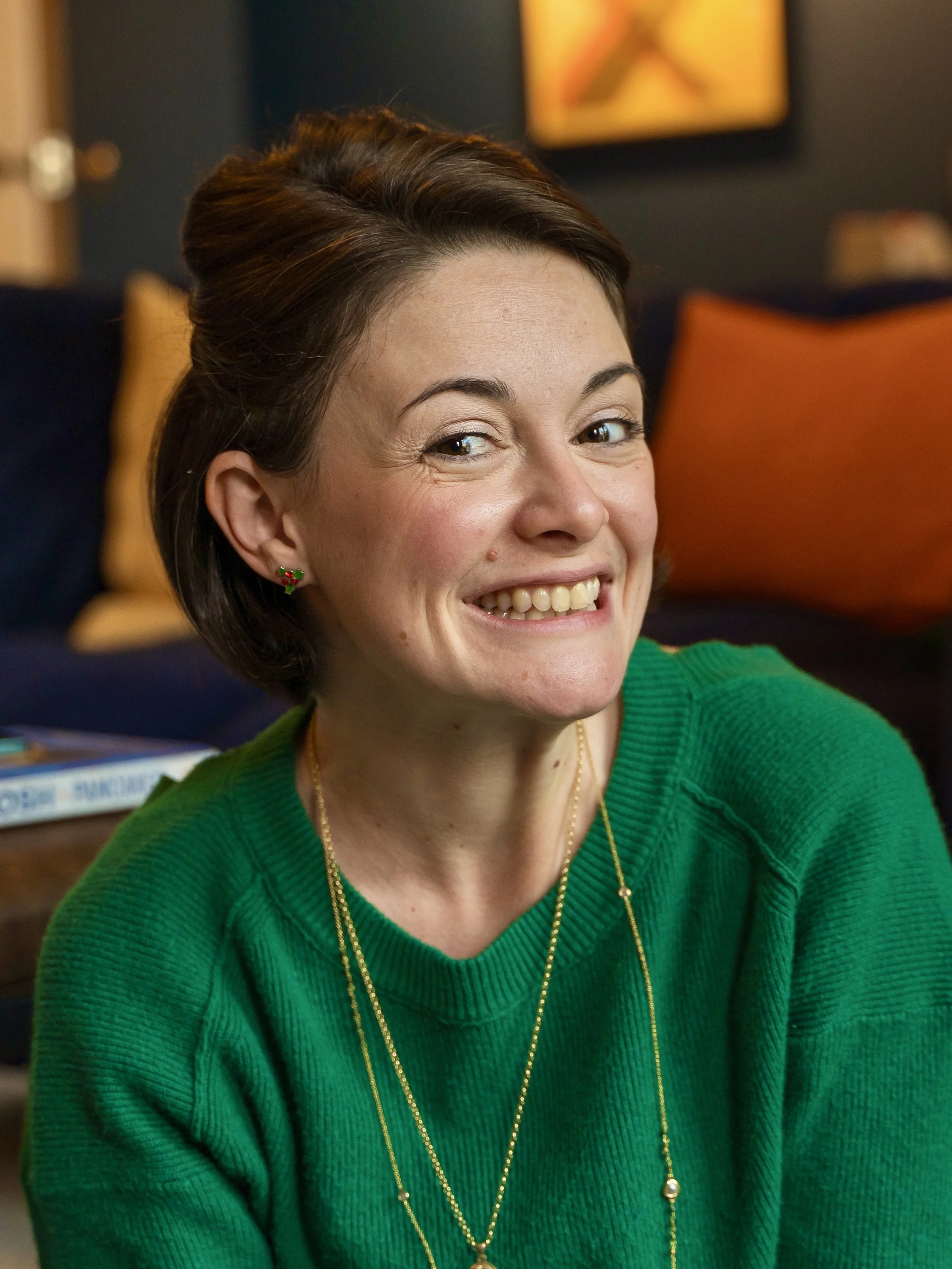 Amie smiling, her head tilted slightly to the right. Her hair is short, half up / half down; she's in a bright green jumper with two, long gold necklaces.