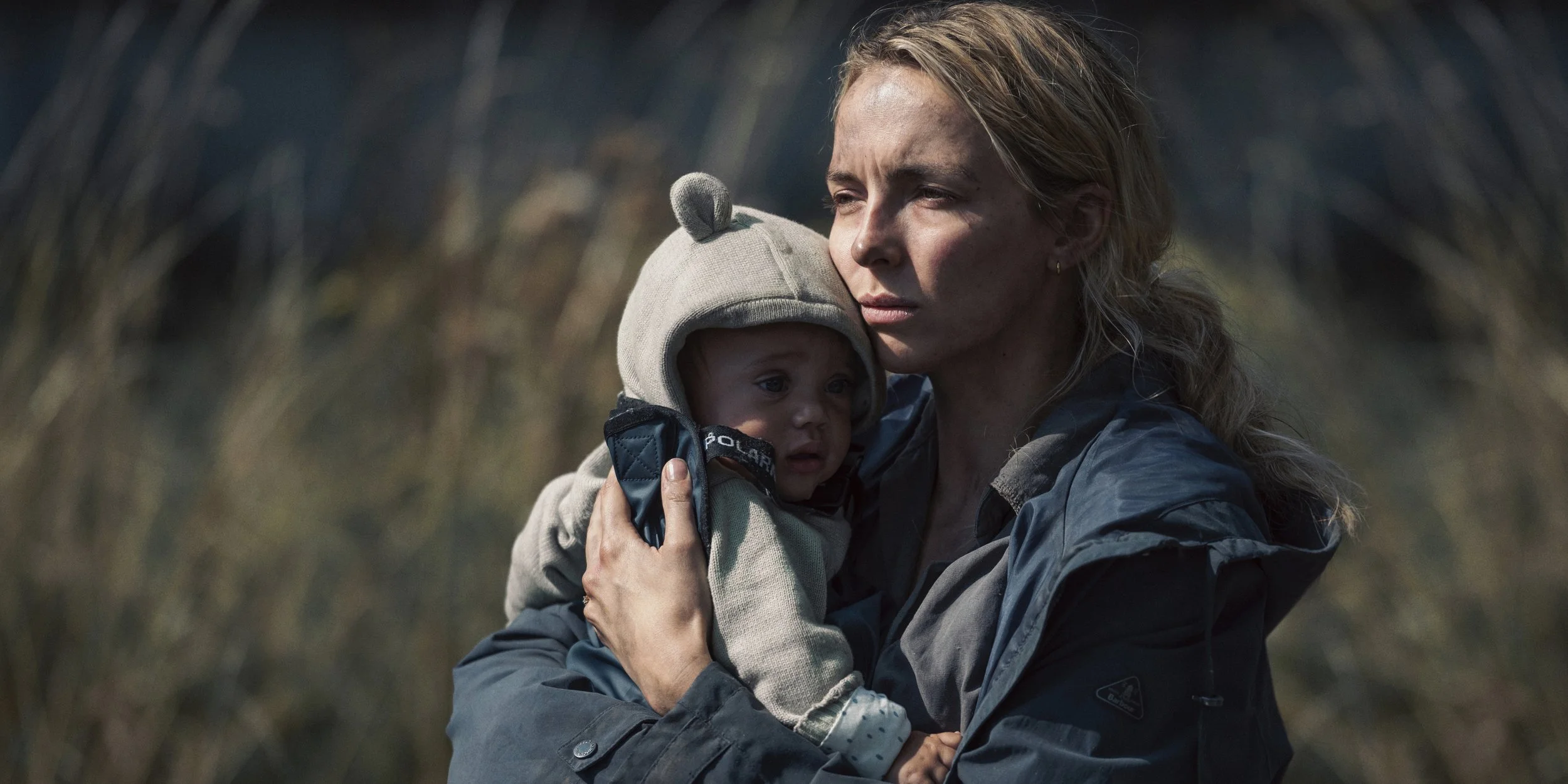 A still from the film adaptation of Jodie Comer wearing a raincoat; she has greasy blonde hair in a ponytail. She's holding a 9 month old baby in a bear suit. She's looking off-camera looking tired and troubled.