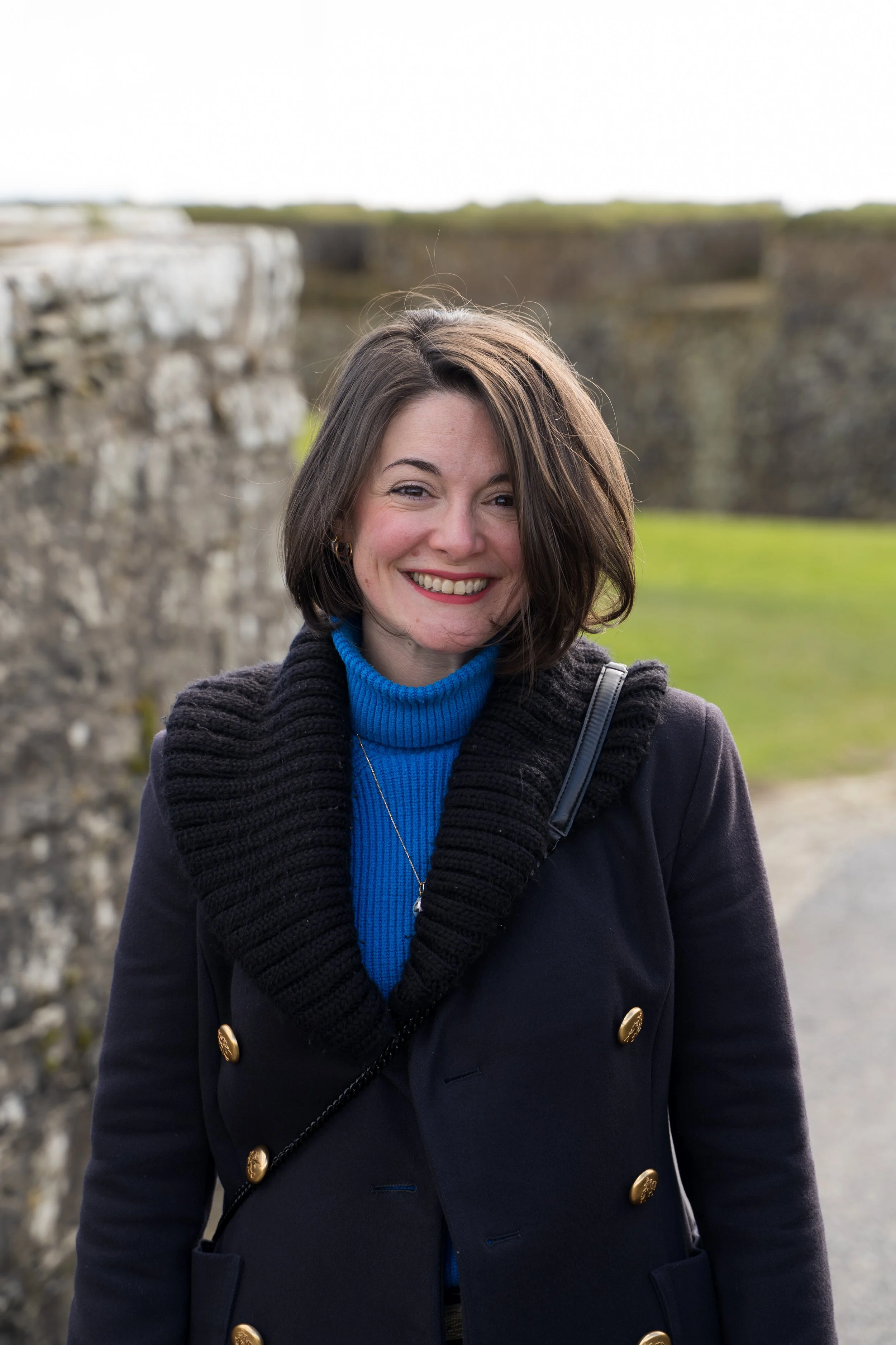 Photo of Amie in front of a stone wall wearing a bright blue turtle neck jumper with a long gold necklace and an open navy peacoat. Her hair is short and she's smiling.
