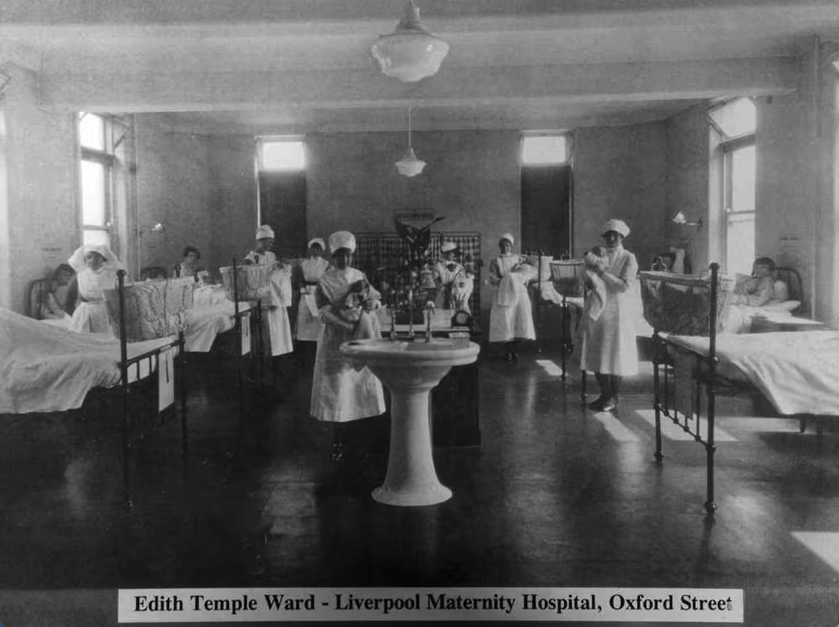 Photo inside a Victorian maternity ward at the old Maternity Hospital in Liverpool. Women in white, floor length nursing uniforms and women in beds on either side of the room.