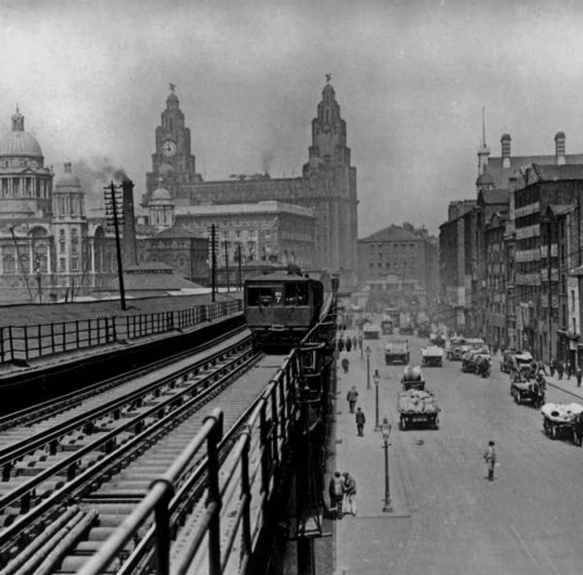 Black and white photo of the Liver building in Liverpool. There's smog and dark clouds in the air and in the foreground is the old Dockland Railway above the street.