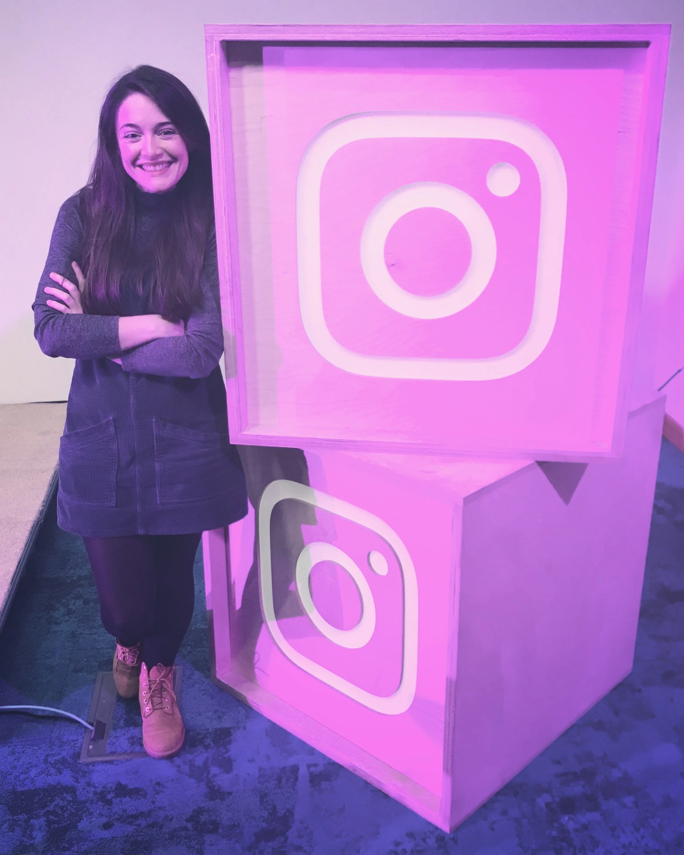 Amie with long hair, smiling, standing with her arms folding as she leans on two big pink blocks with the Instagram logo embossed on them.