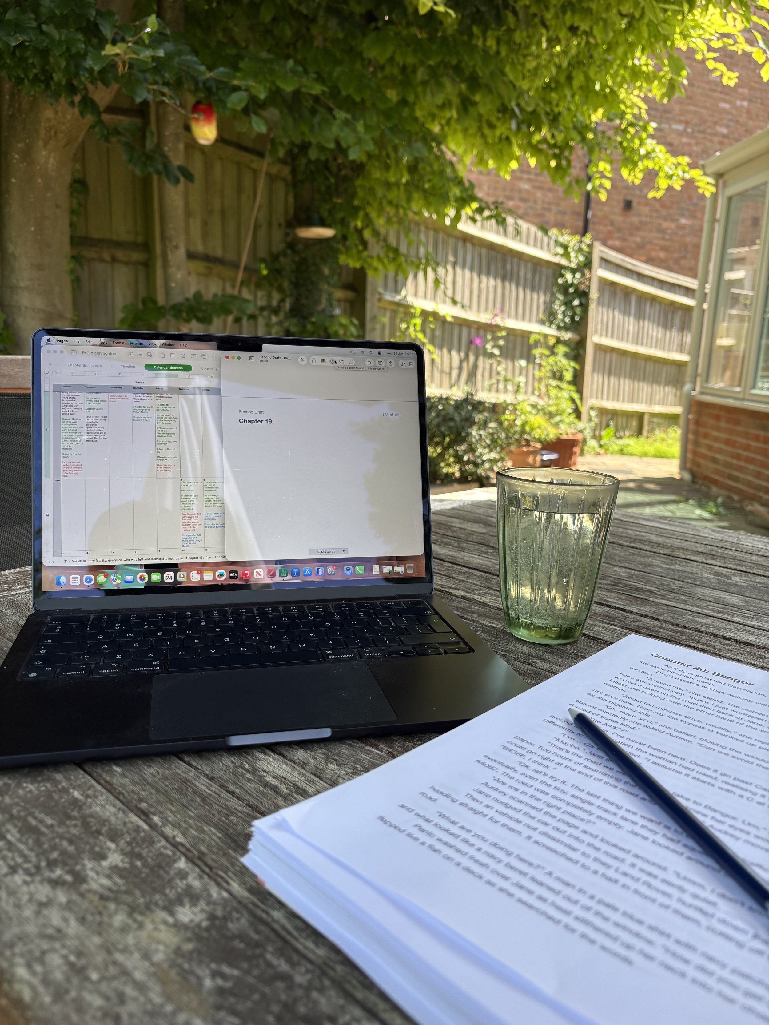 On an outdoor table in the shade of a garden there's a printed manuscript and pencil beside an open laptop; on the screen is a spreadsheet (unreadable) and an open, blank doc with "Chapter 19" at the top.