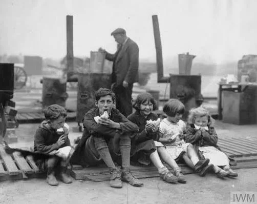 A group of four children (approx age 3-12) sit on a curb in war-torn Liverpool eating sandwiches. One girl is laughing but the oldest boy stares straight at the camera.
