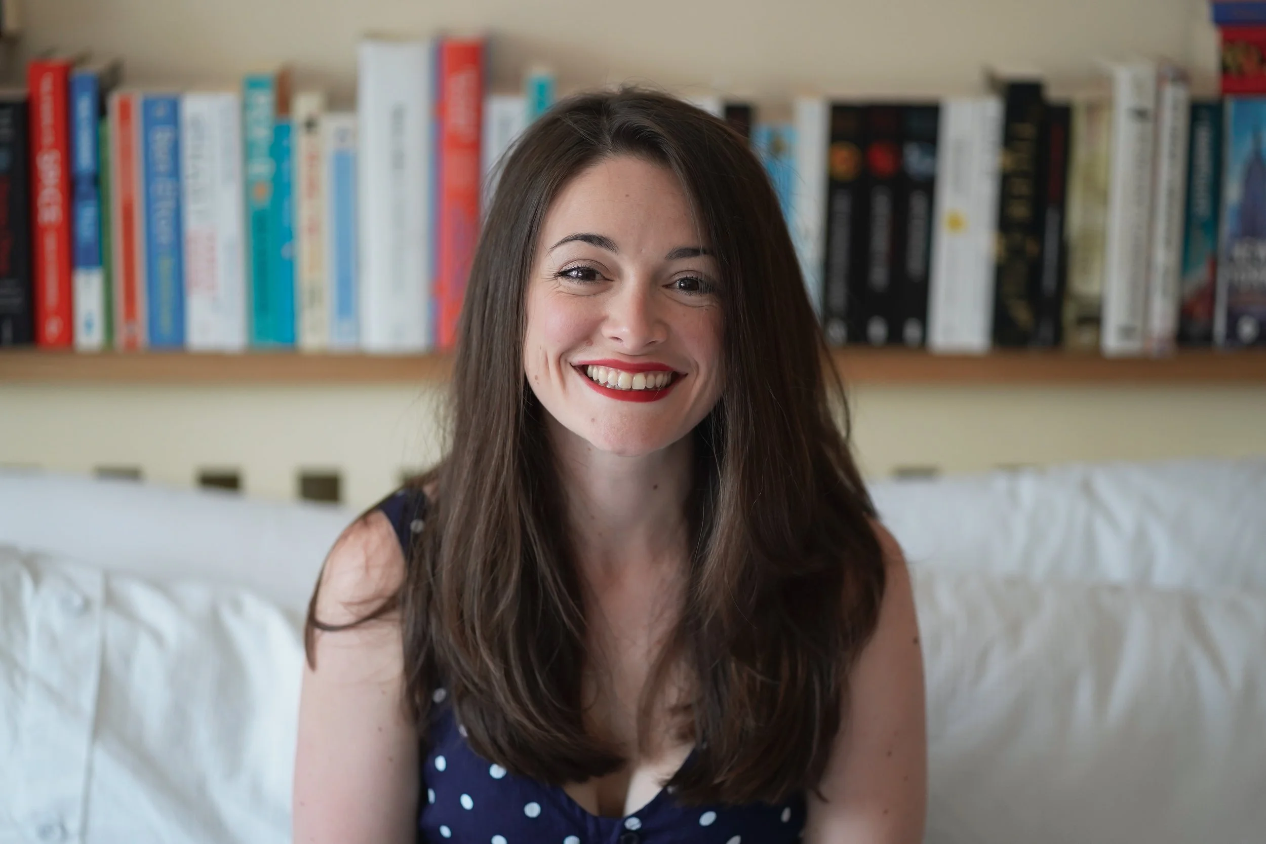 Photo of Amie (long brown hair, smiling with red lipstick) in a navy and white polkadot top sitting in front of a single shelf of books.