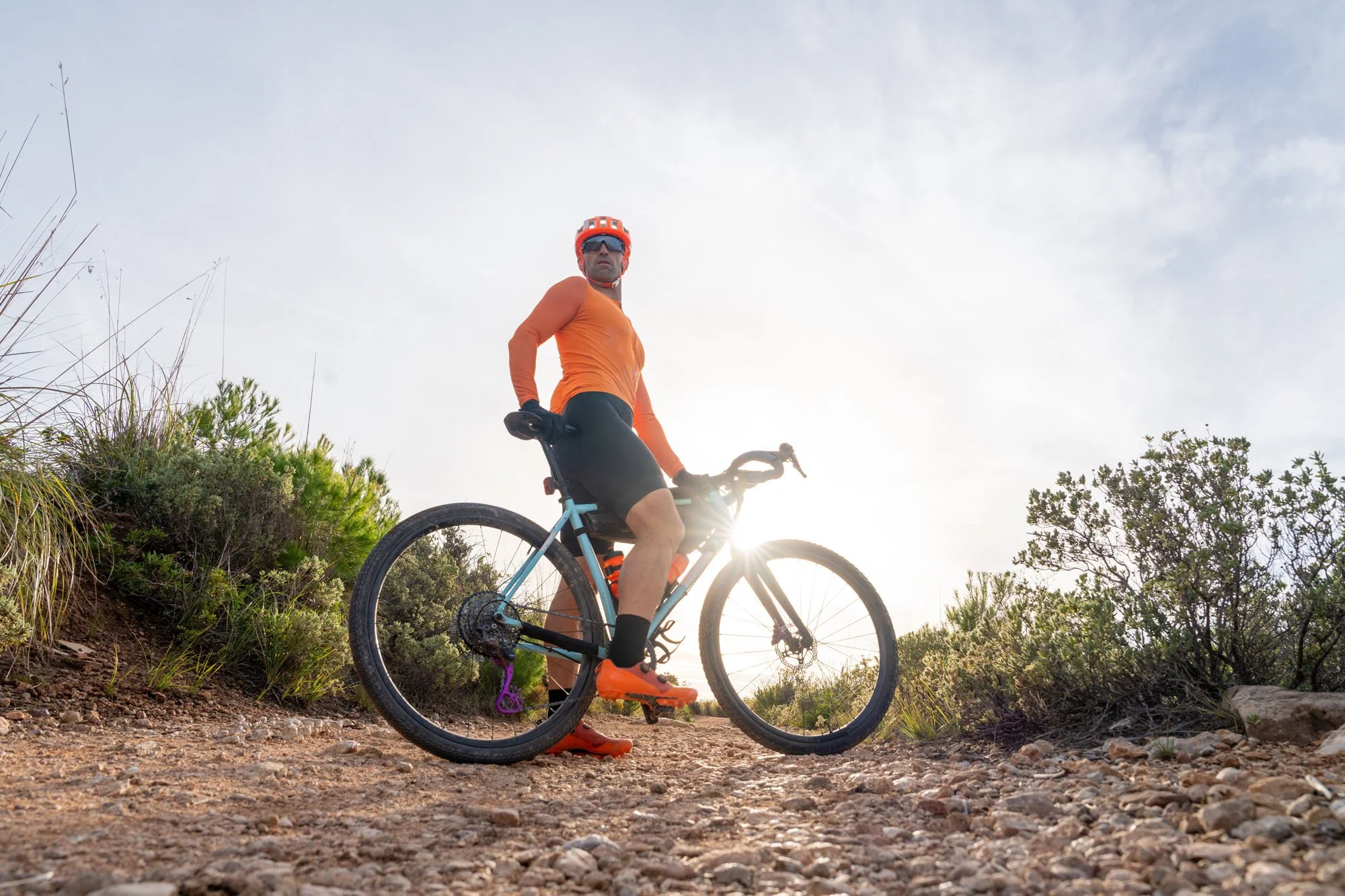 Hombre usando casco y gafas de sol montando en bicicleta por un sendero natural durante la tarde, con arbustos y árboles en el fondo.