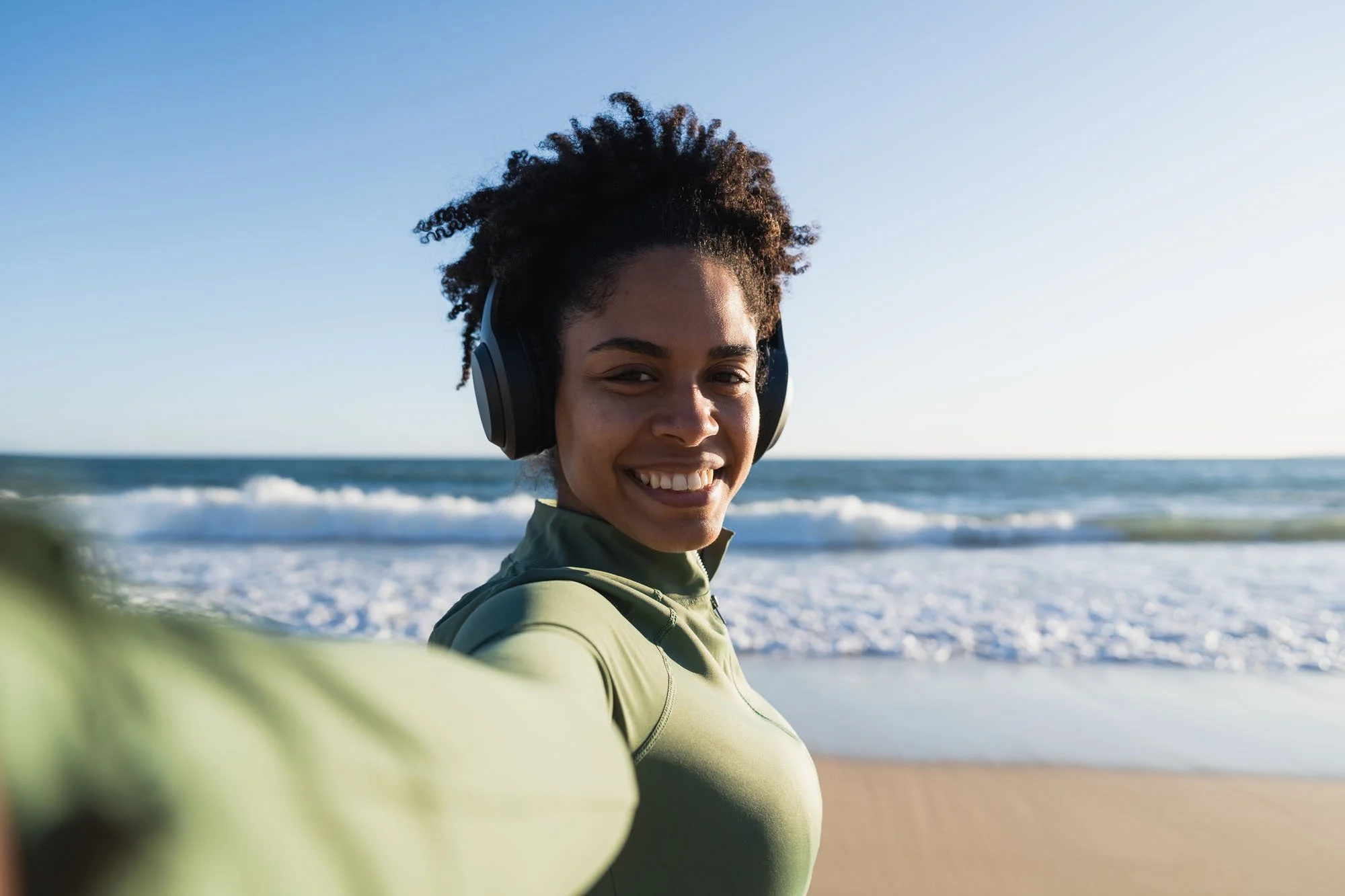 Joven mujer con auriculares en la playa, sonriendo, con el mar y el cielo despejado al fondo.