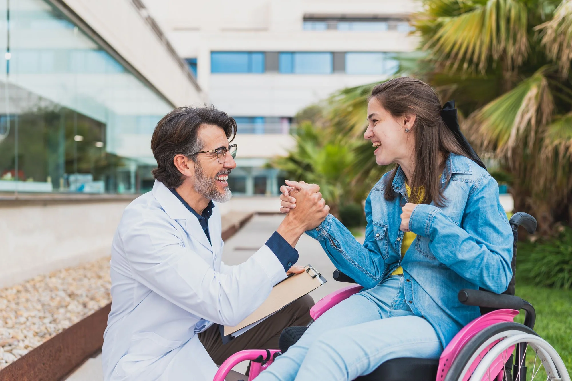 Doctor y joven en silla de ruedas dándose la mano y sonriendo en un parque frente a un edificio moderno.