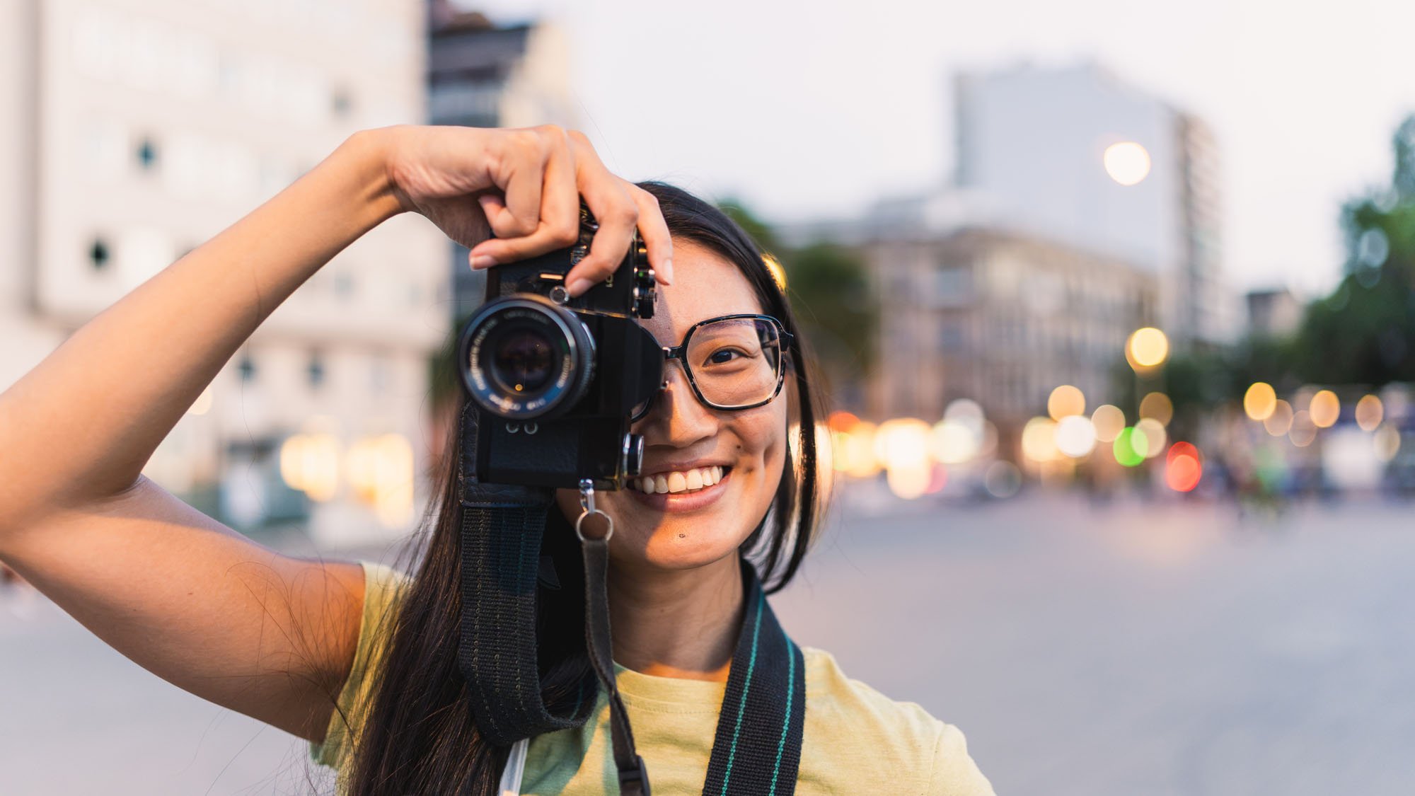Joven mujer sonriendo y tomando una fotografía con una cámara en una calle de la ciudad al atardecer.
